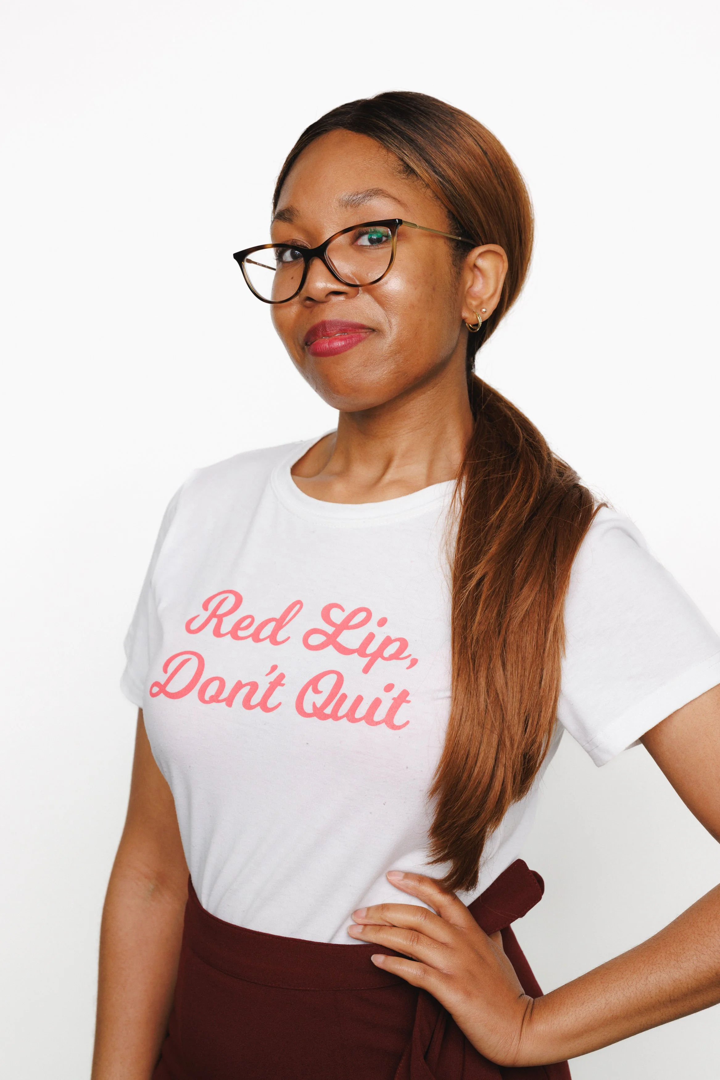 A woman with long brown hair, glasses, and earrings, wearing a white t-shirt with pink text that reads 'Red Lip, Don't Quit,' and maroon pants, standing against a white background.