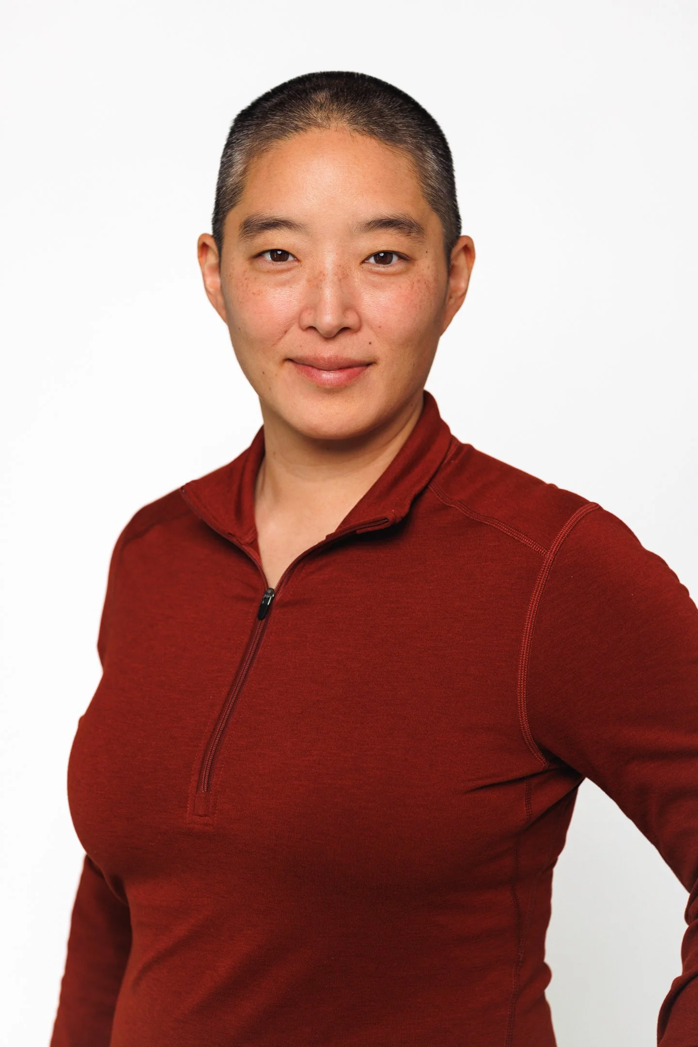 A woman with a bald head and light skin, wearing a maroon zip-up athletic shirt, smiling at the camera against a plain white background.