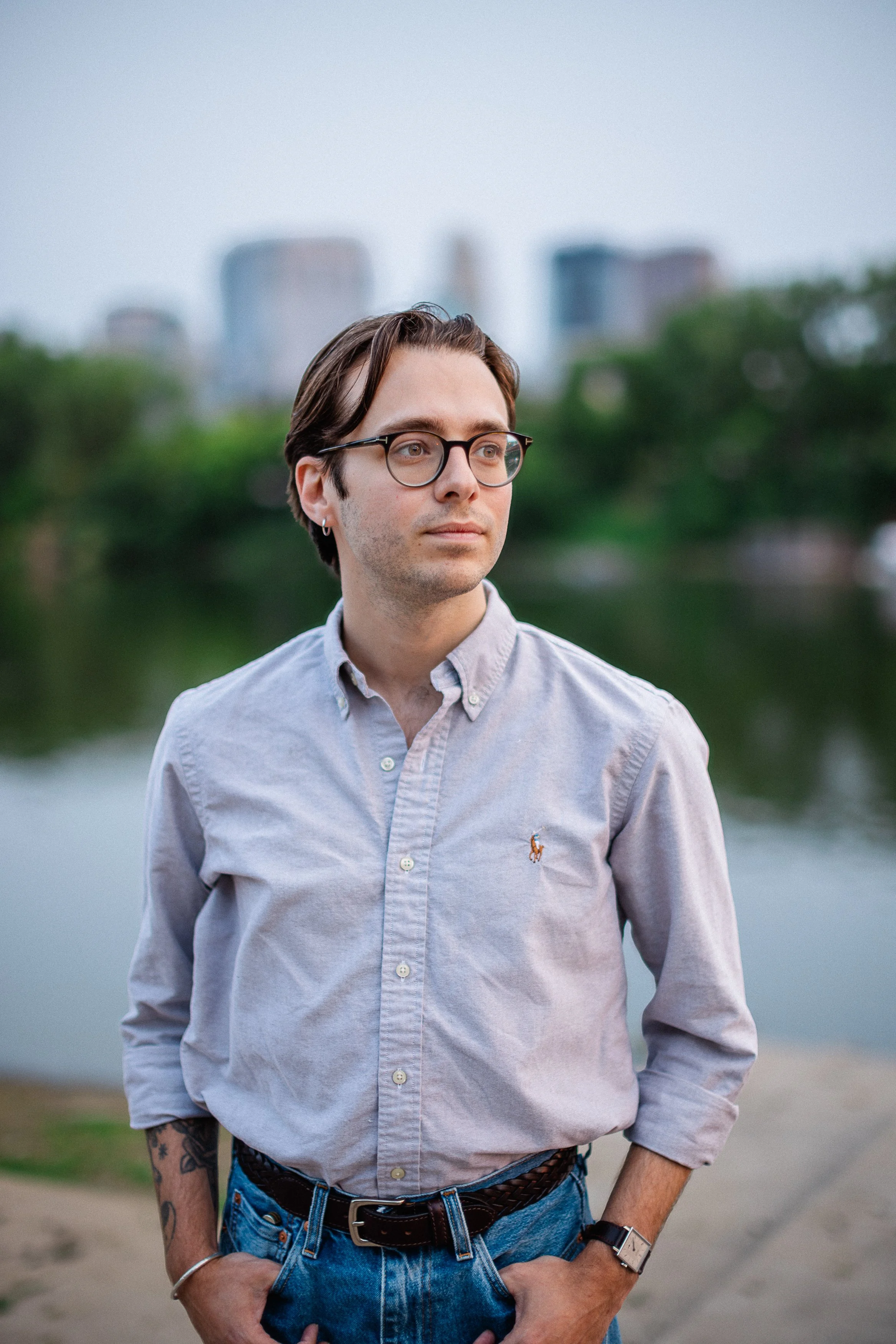 A young man with glasses and a tattoo on his arm wearing a light gray button-up shirt and blue jeans, standing outdoors near a body of water with a cityscape in the background.