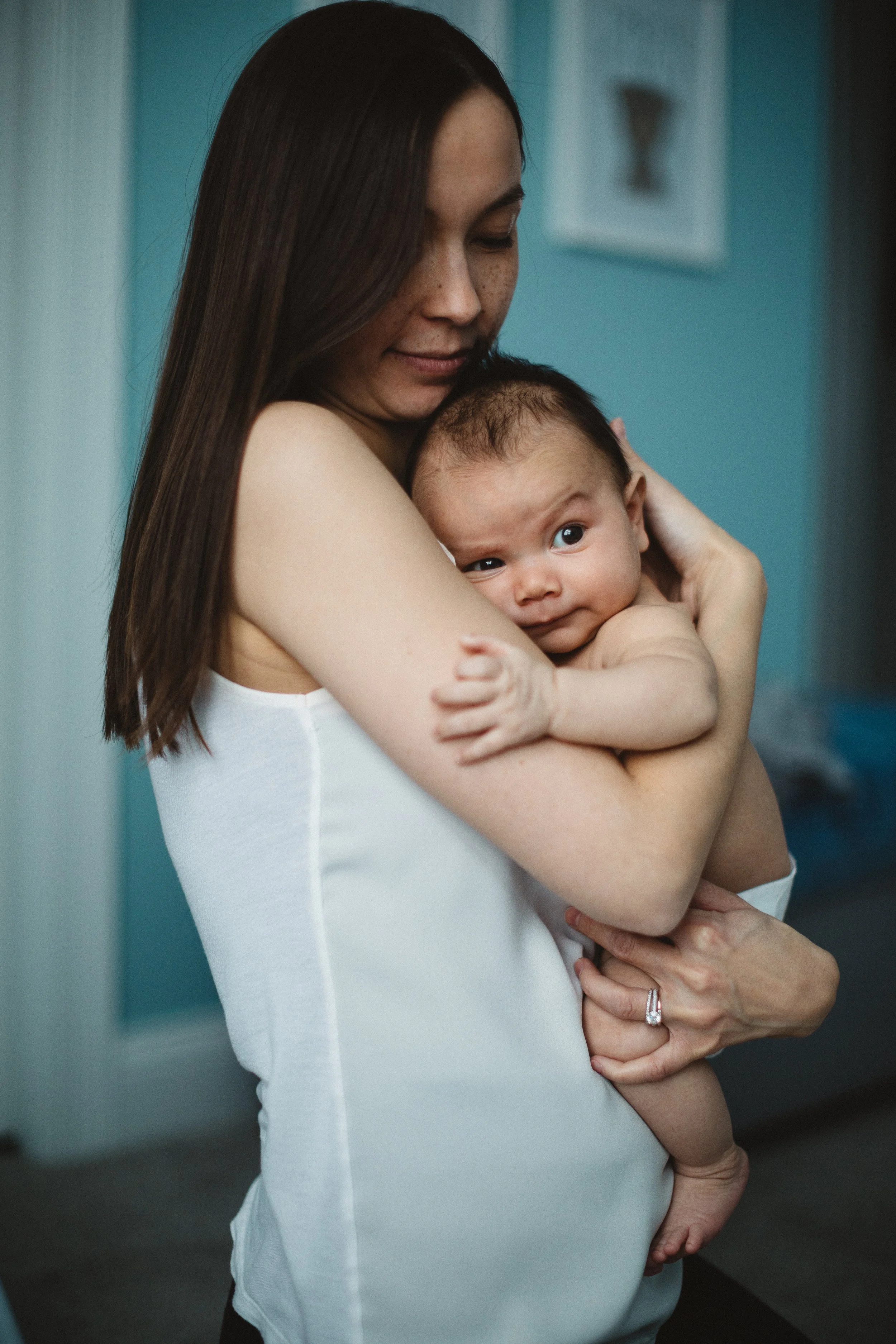 A woman holding a baby close to her chest indoors, with a blue wall in the background.