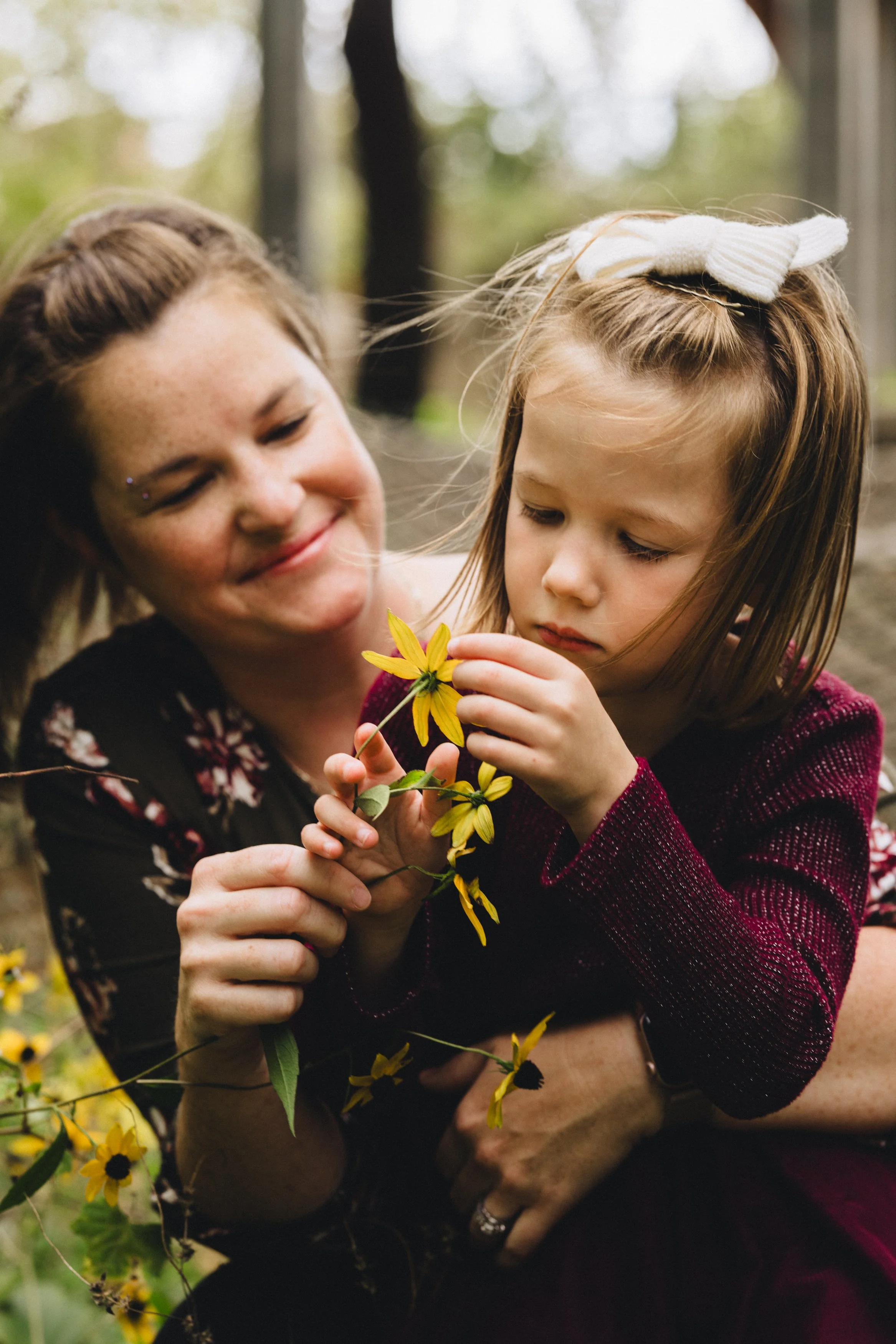 A woman and a young girl outdoors, closely examining yellow flowers. The woman is smiling, and the girl appears focused on the flower she is holding.