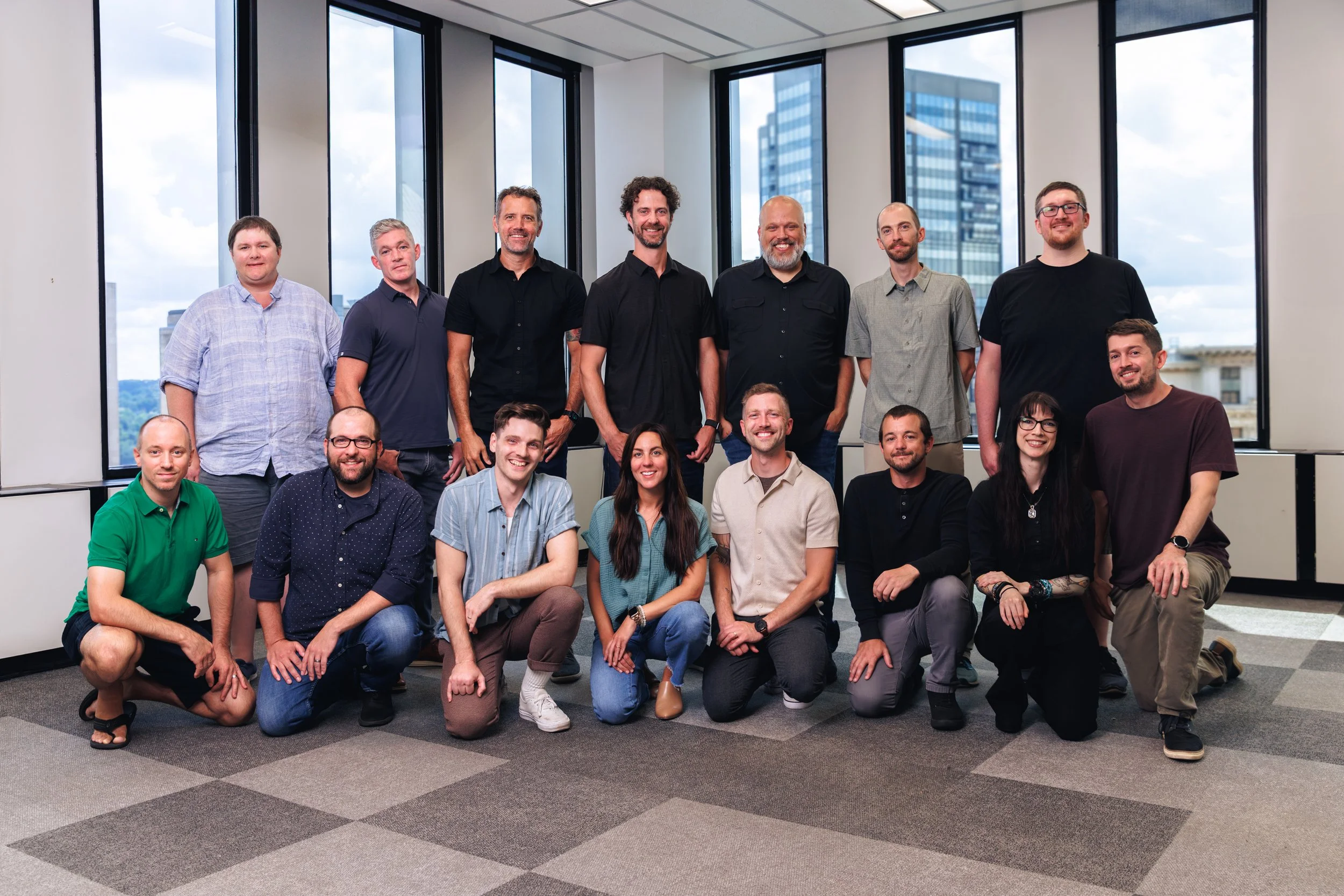 Group photo of fifteen diverse adults, including men and women, posing together in a modern office space with large windows and city views in the background.