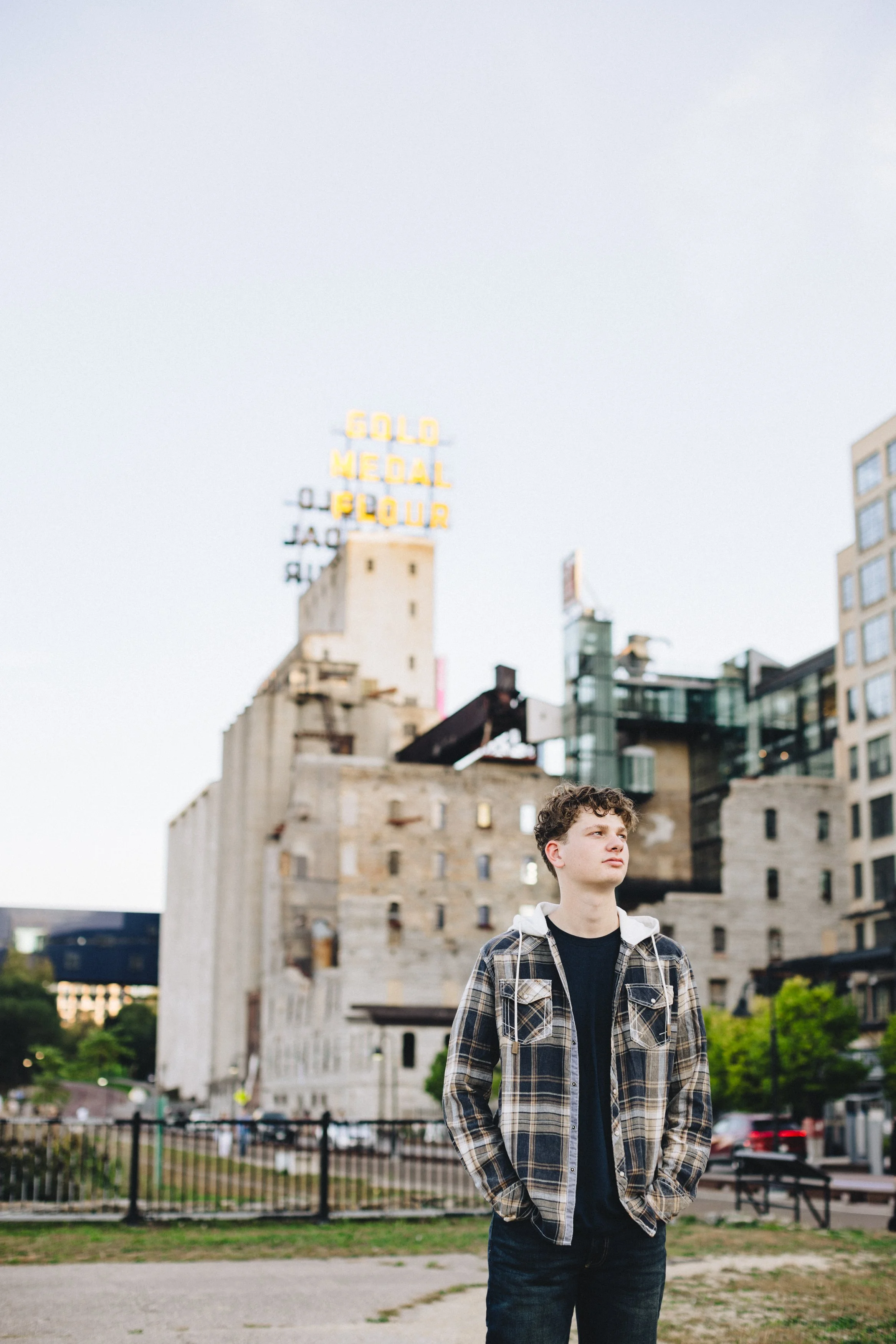 A young man with curly hair and a plaid jacket stands outdoors with hands in pockets, looking off into the distance, with a city landscape and a sign reading 'Gold Medal Flour' in the background.