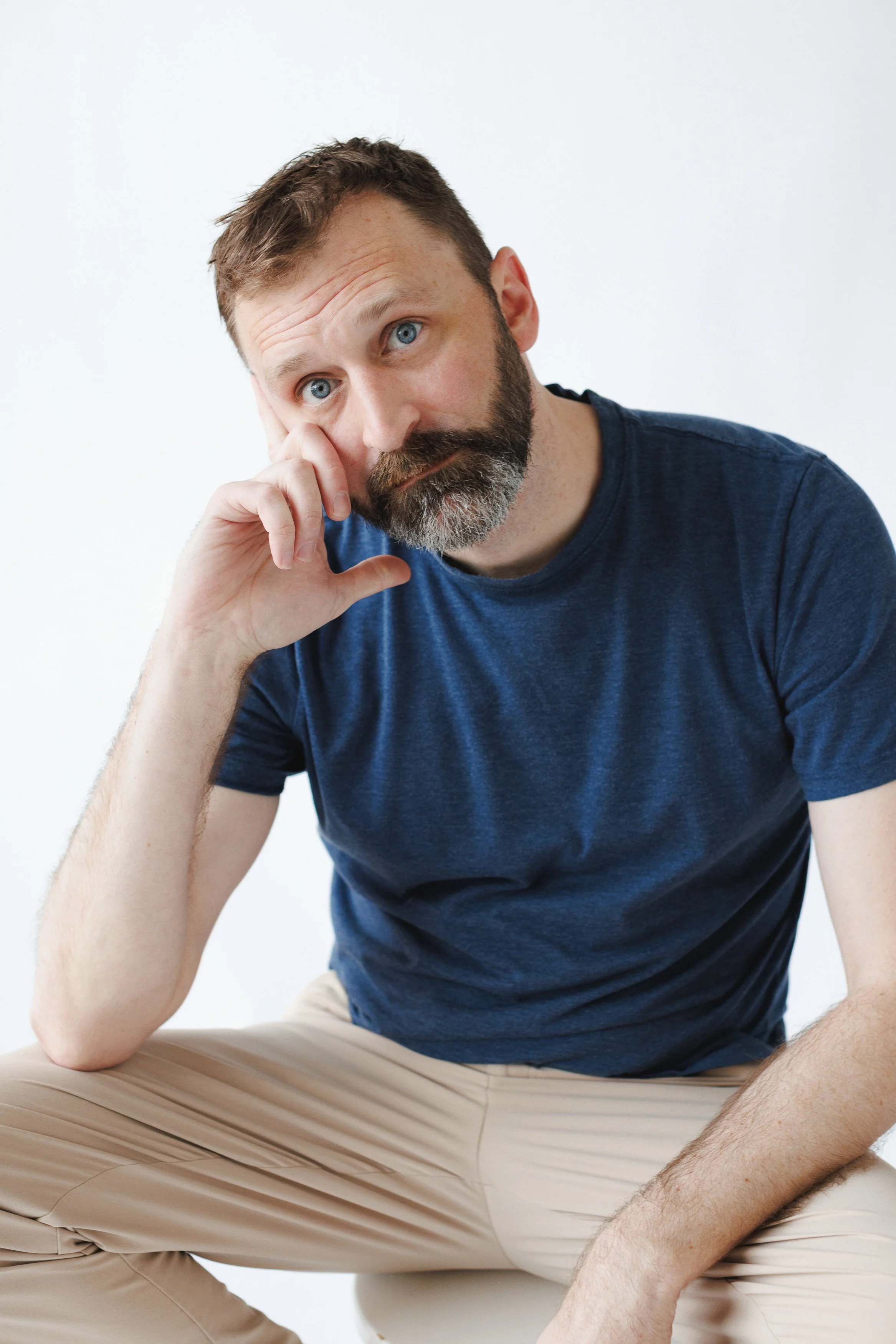 A man with a beard and blue eyes, wearing a blue t-shirt and beige pants, sitting against a plain white background, resting his head on his hand with a pensive expression.