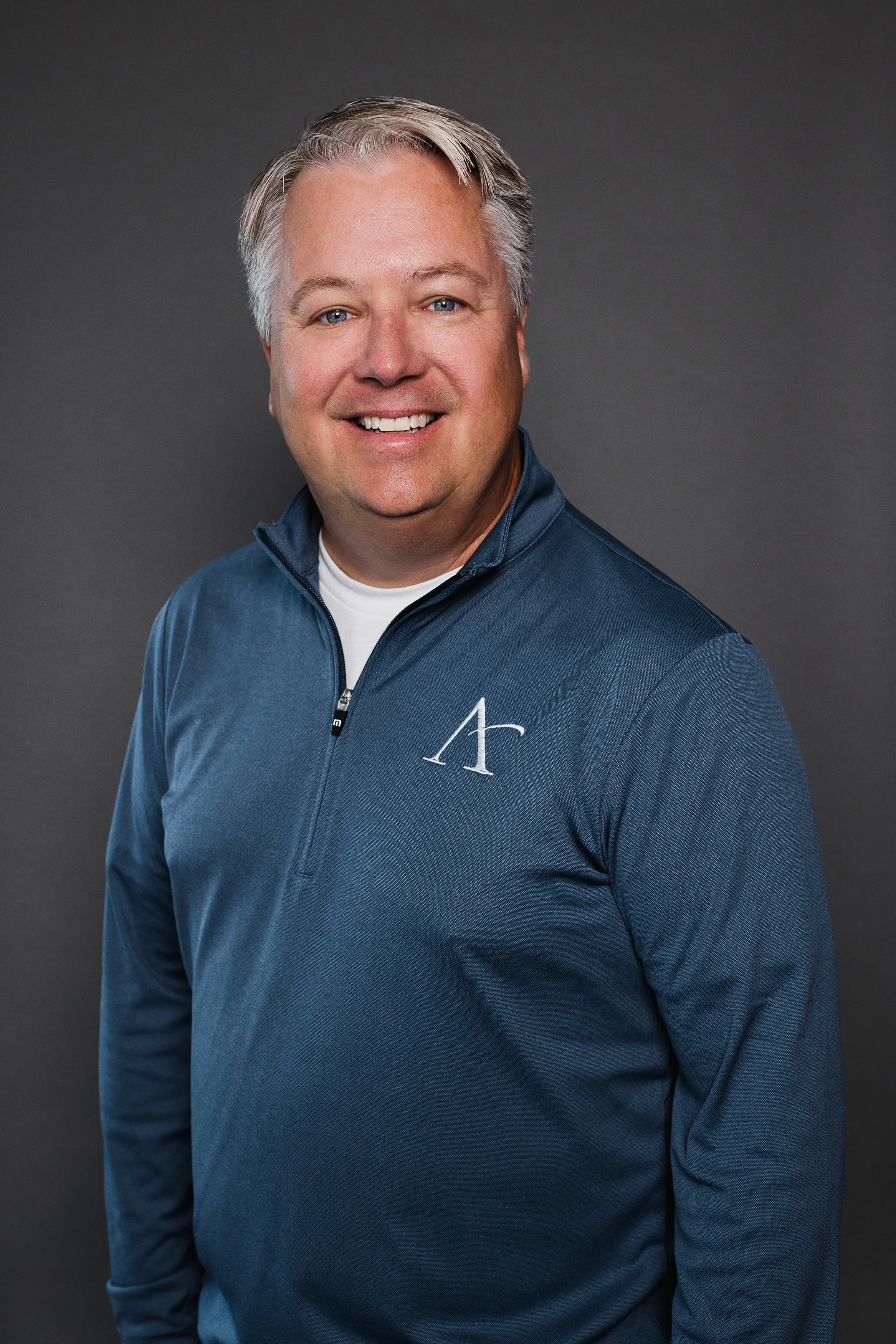 A smiling middle-aged man with gray hair and blue eyes wearing a navy blue quarter-zip sweater with a white logo on the left chest, standing against a dark gray background.