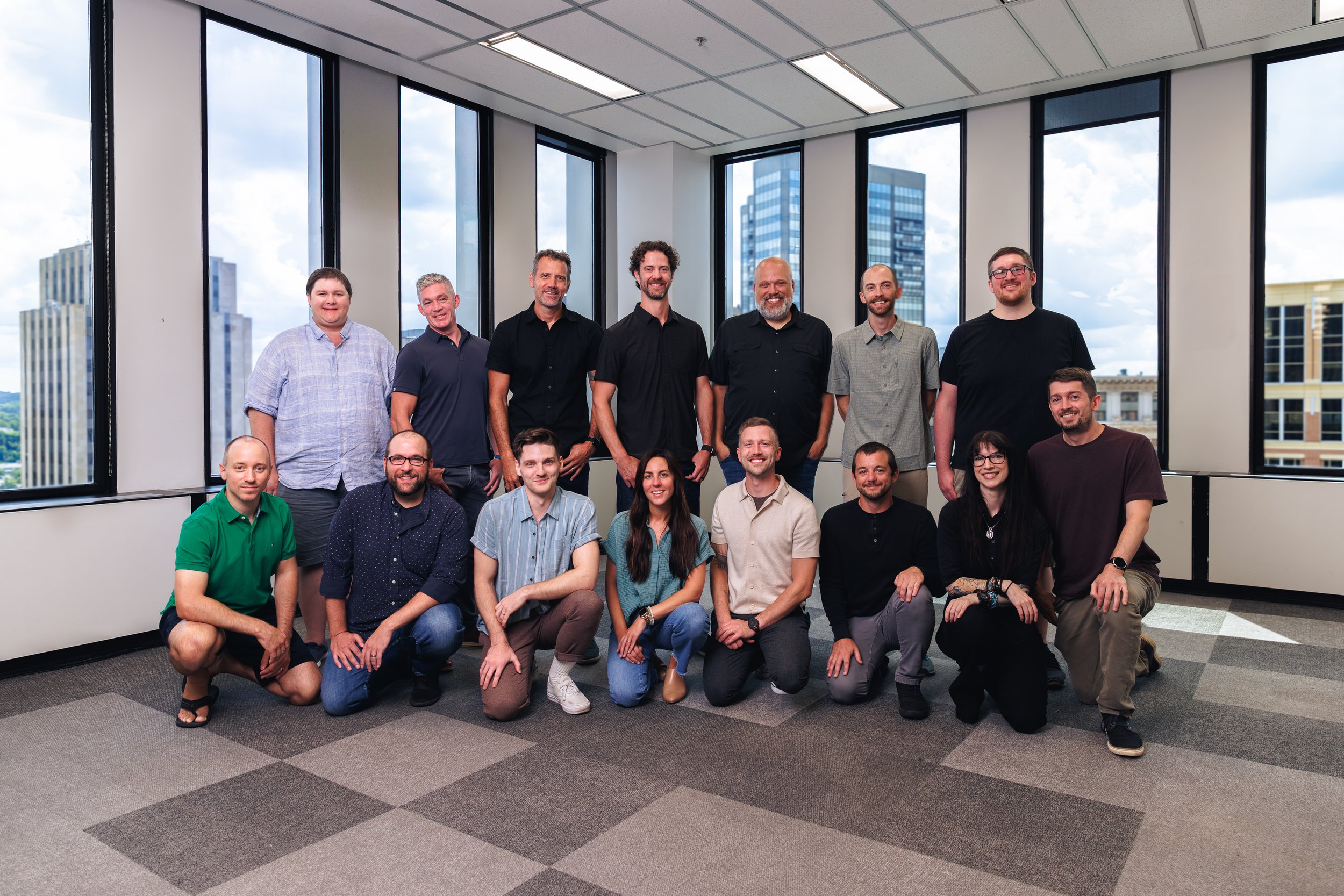 Group of 15 people posing in an office with large windows and city view.