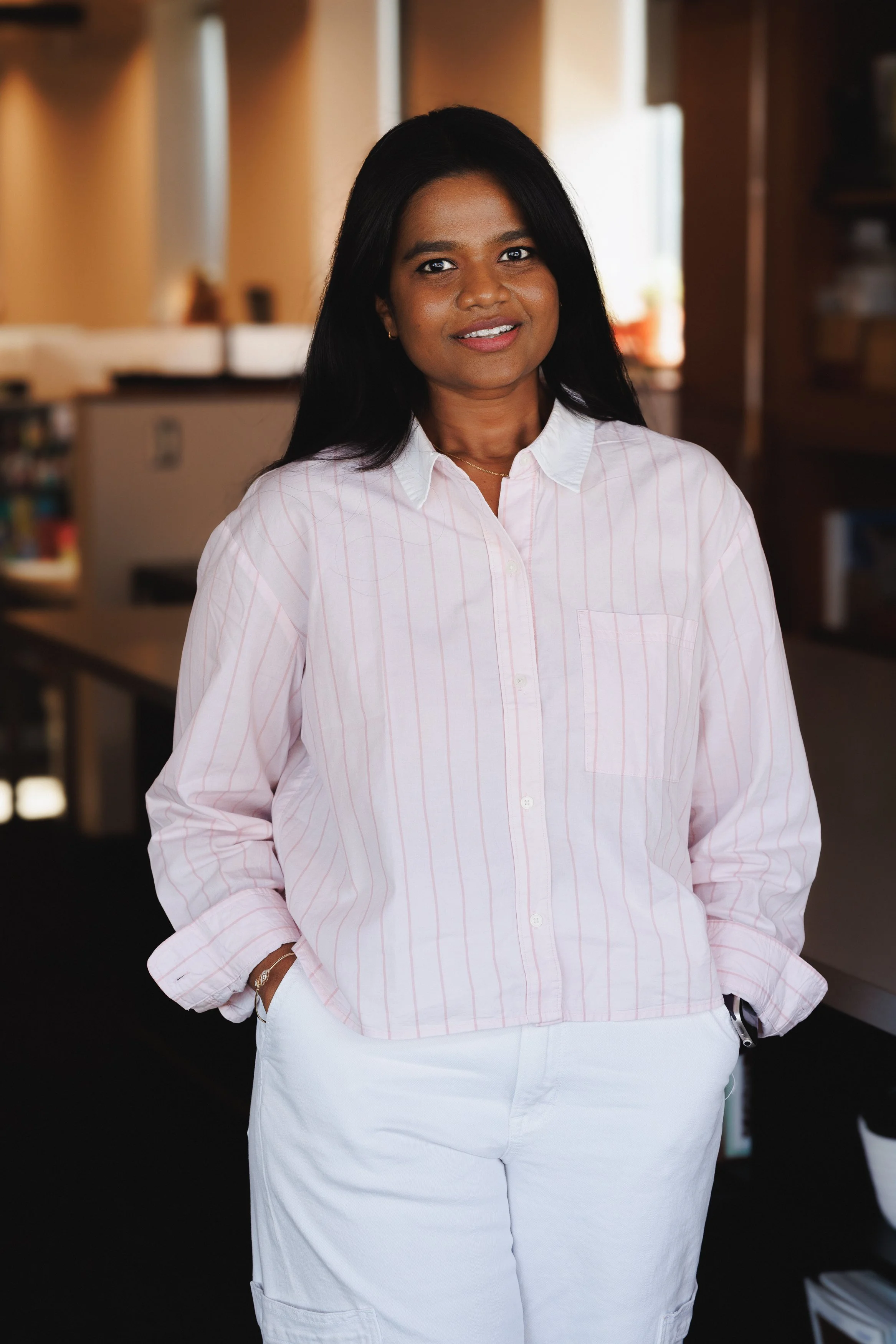 A woman with long dark hair wearing a pink striped button-up shirt and white pants, standing in an indoor setting with a blurred background.