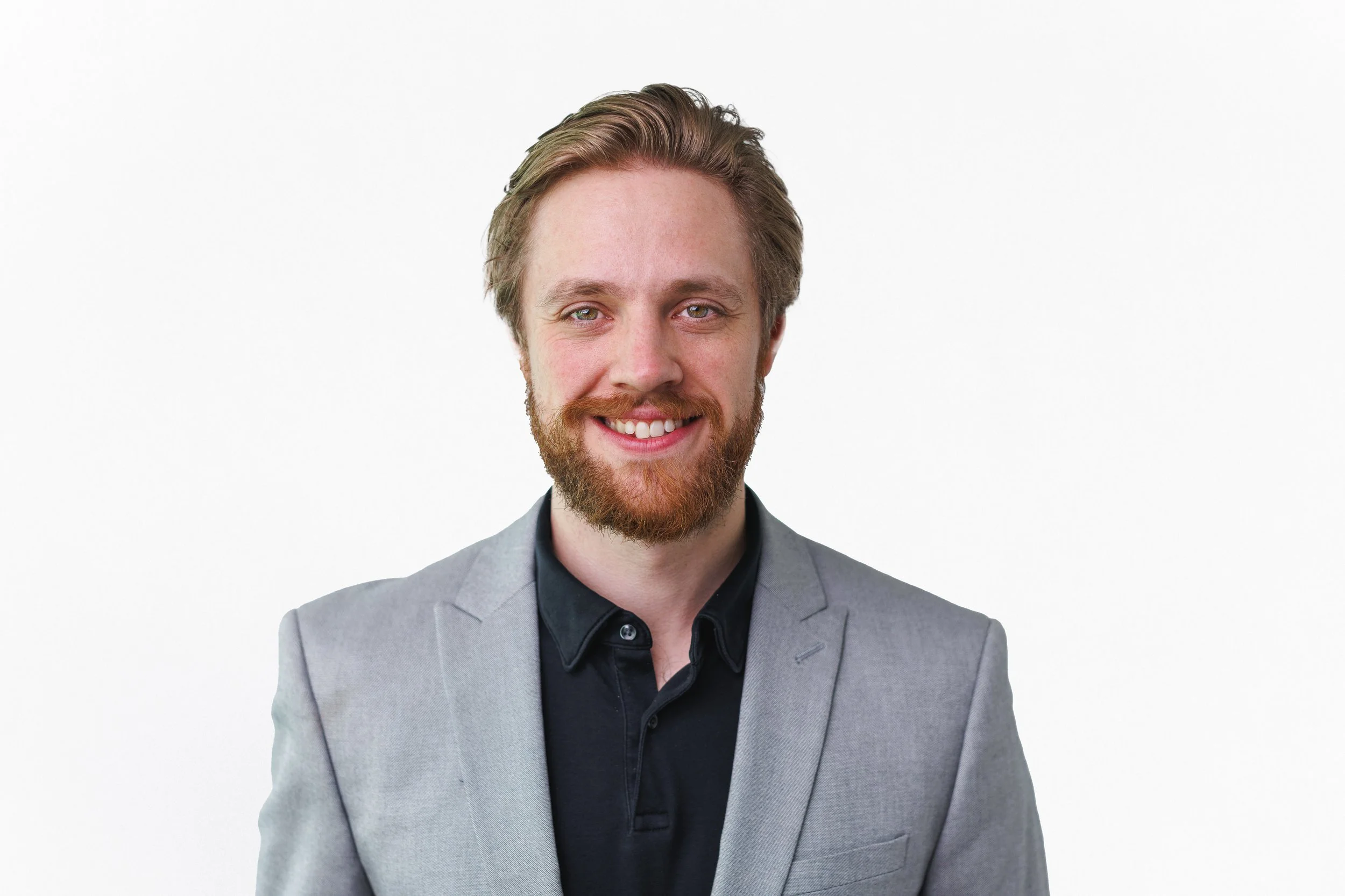 Headshot of a young man with a beard and blond hair, smiling, wearing a gray blazer and black shirt against a white background.