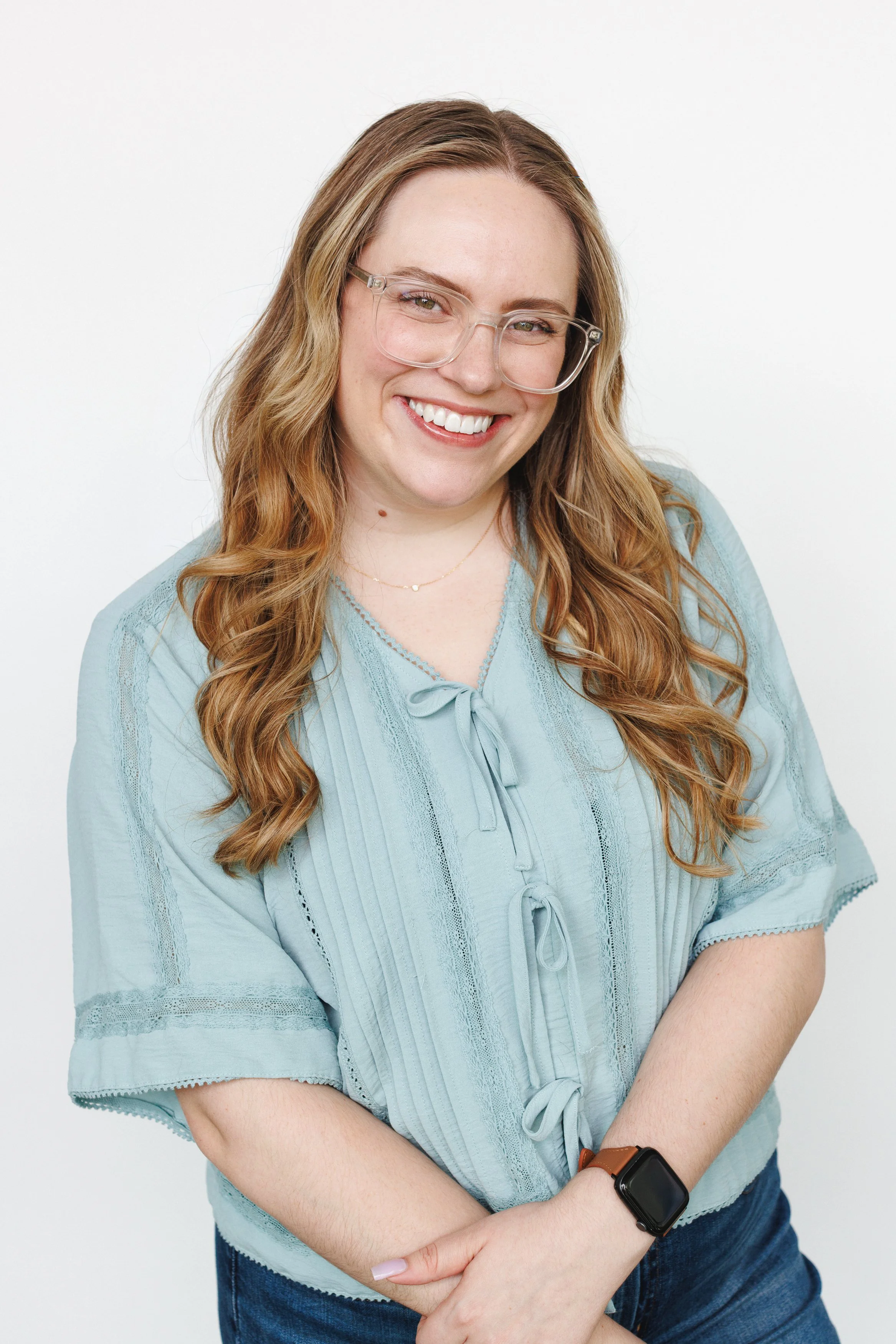 A woman with long wavy red hair, wearing glasses, a light blue blouse with bows, and a smartwatch, smiling at the camera against a plain white background.