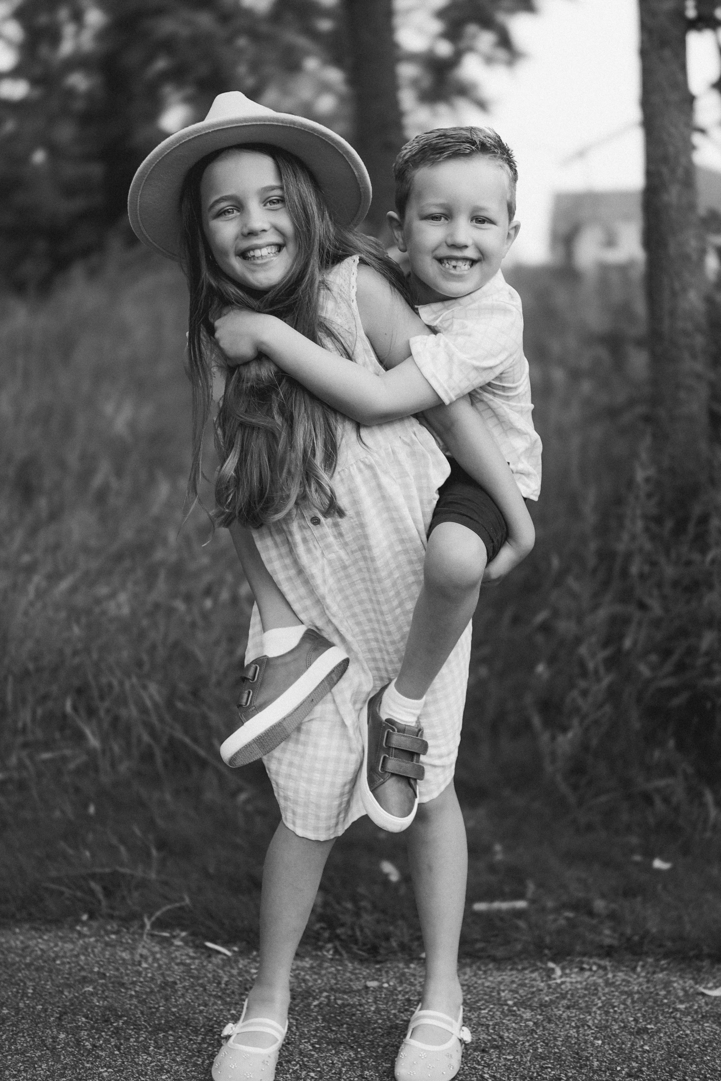 A young girl wearing a hat and a checkered dress is being carried on a boy's back, both smiling and embracing outdoors in a grassy area with trees in the background.