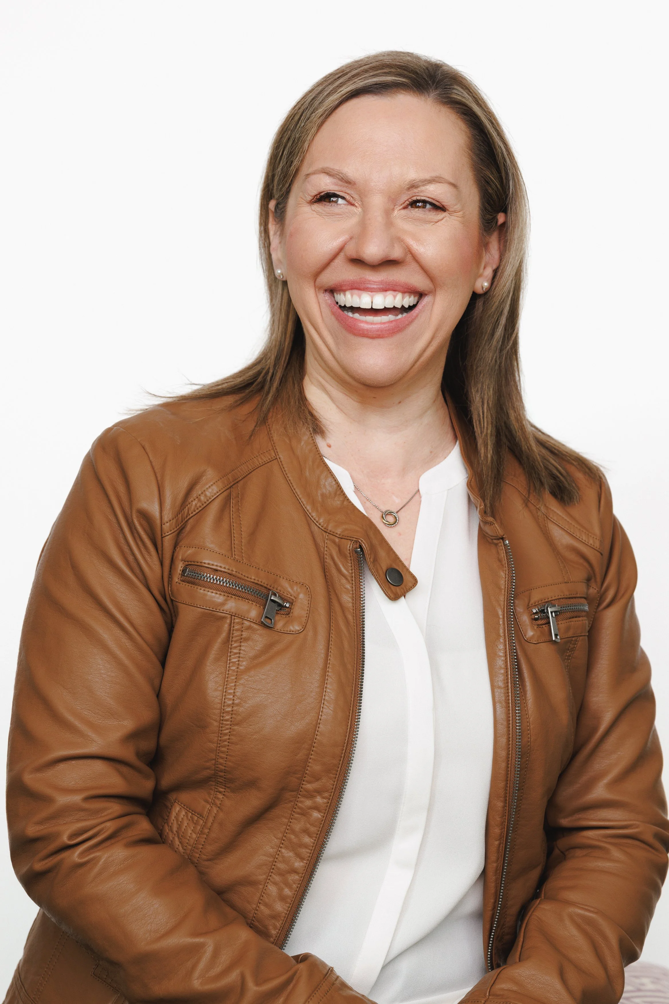 A woman with shoulder-length light brown hair, wearing a brown leather jacket over a white shirt, smiling and looking to her right against a plain white background.