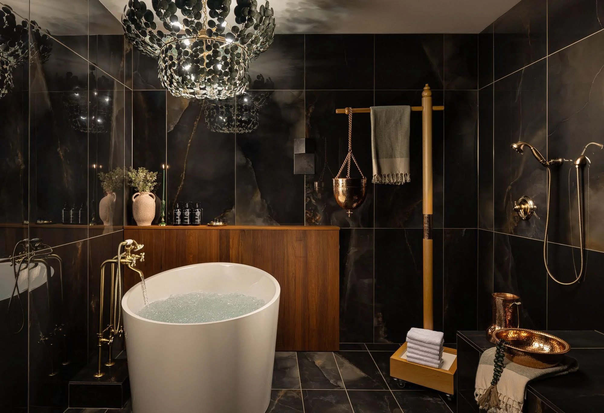 Luxurious bathroom with black marble walls, a white freestanding bathtub, gold fixtures, a dark wood vanity, decorative plants, and a statement chandelier.
