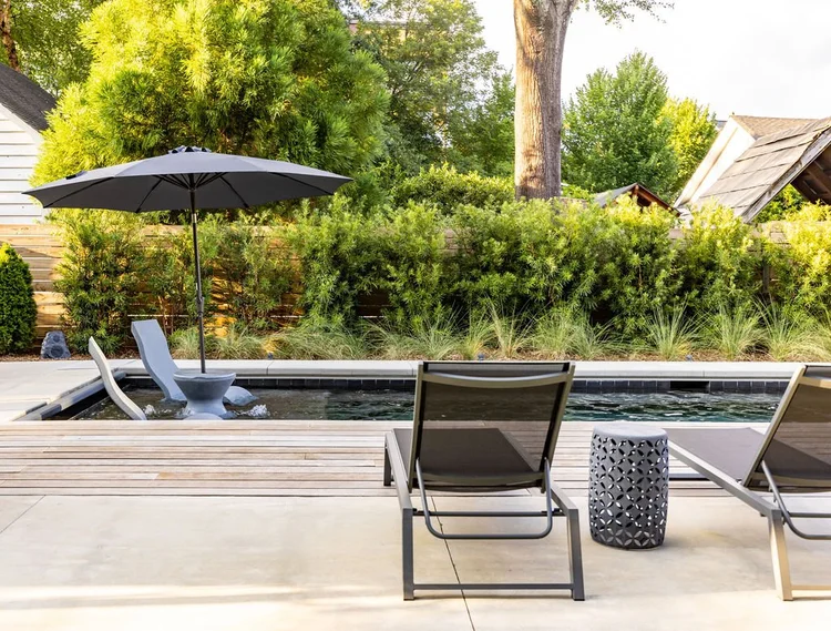 Backyard pool area with two lounge chairs and a small table, surrounded by lush greenery and a wooden fence, with umbrellas and garden structures in the background.