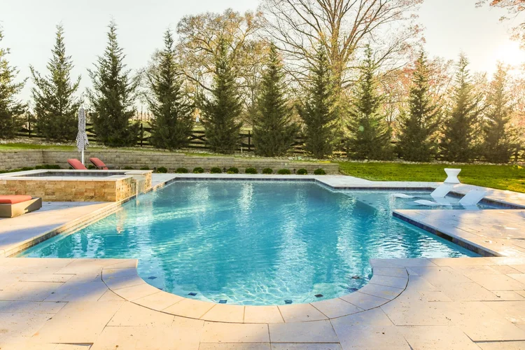 Inground swimming pool with lounge chairs, surrounded by trees, during late afternoon or early evening sunlight.