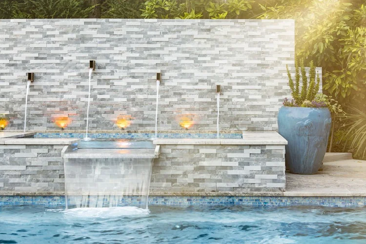 Outdoor water fountain feature with multiple streams of water flowing from spouts in a gray stone wall behind a swimming pool, with a large blue pot containing green plants and purple flowers on the side, and lush greenery in the background.