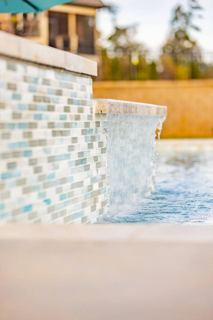 Close-up of a water feature with water cascading over a tiled edge, with a pool below, and a wooden fence and trees in the background.