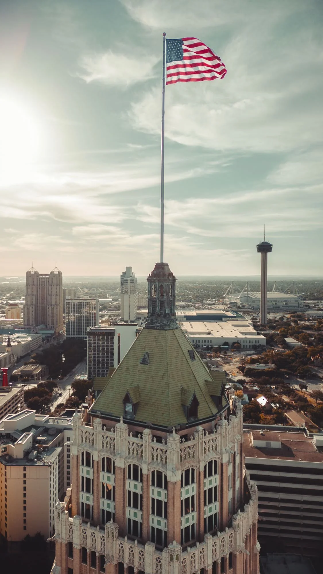 An American flag flying high atop a historic building in a cityscape, with modern skyscrapers and a tower in the background under a partly cloudy sky.