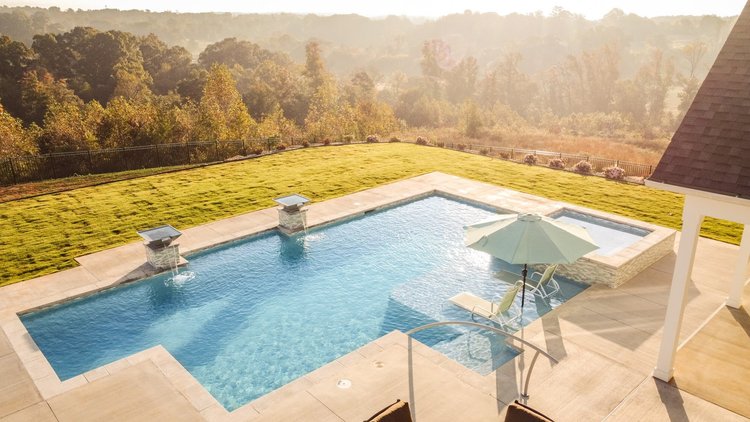 Backyard with swimming pool, lounge chairs, patio umbrella, and fountain, surrounded by green lawn, trees, and distant hills.