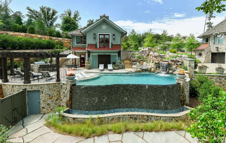 Luxury backyard with an in-ground swimming pool featuring a waterfall, surrounded by rocks, potted plants, lounge chairs, and a two-story house with blue and stone exterior.