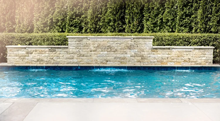 A swimming pool with a stone wall feature that has small water spouts, surrounded by a hedge and tall trees in the background.