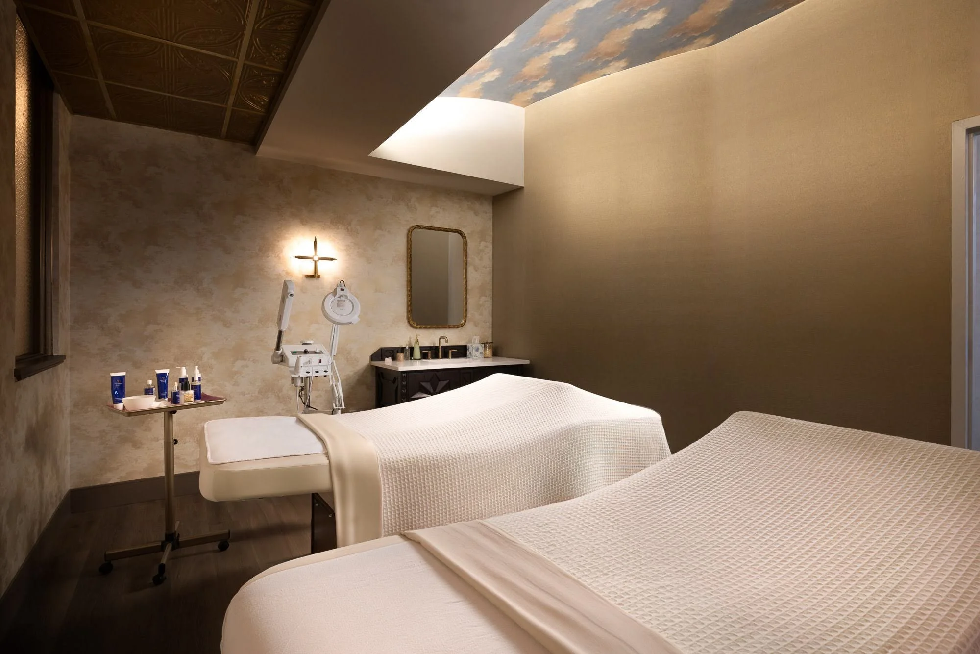 Spa treatment room with two massage beds covered in white sheets, a wall mirror, a lamp, and skincare products on a nearby table, with a ceiling mural of clouds.