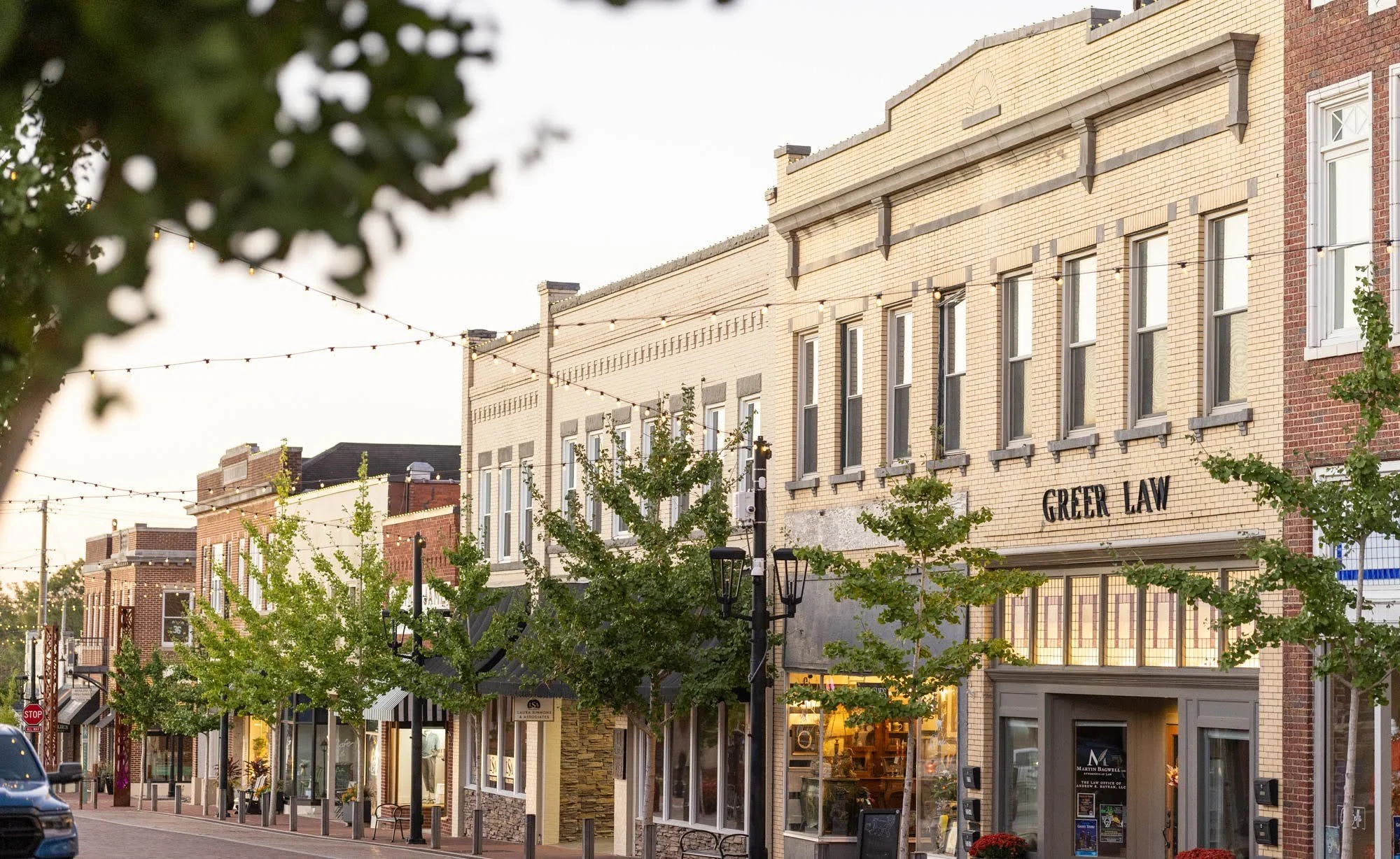 Street view of a downtown area with brick storefronts, trees, and string lights overhead, including a building with a sign that reads 'GREER LAW'.