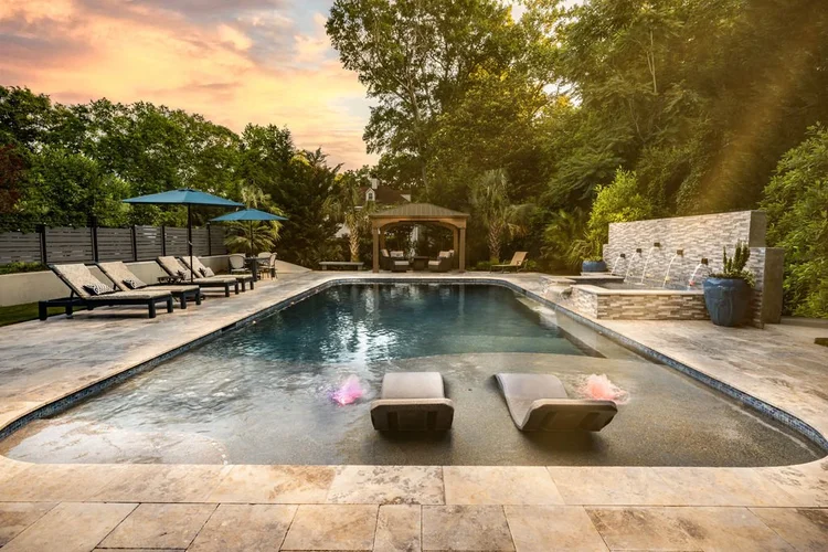 Backyard pool area with lounge chairs, umbrellas, and a waterfall feature surrounded by trees and greenery at sunset.