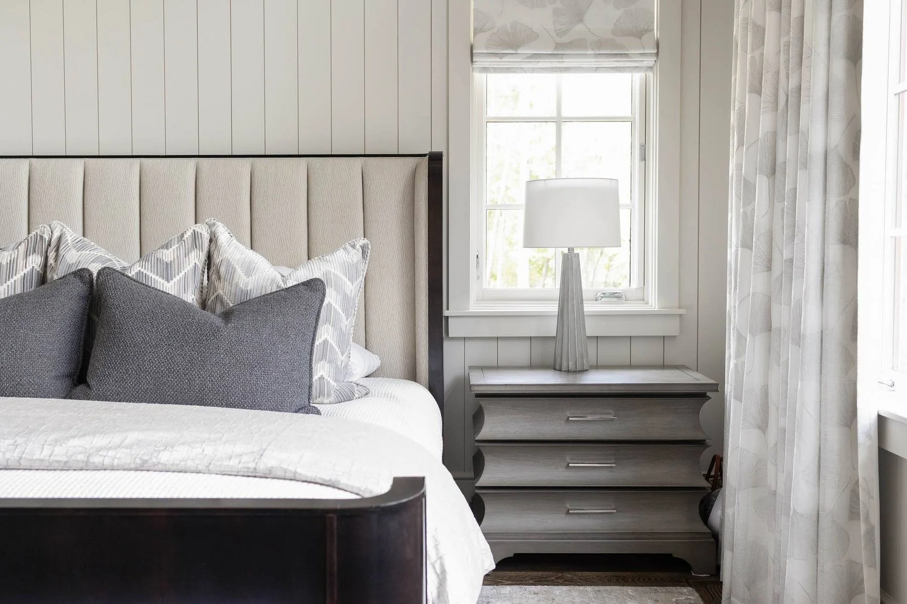 A bedroom with a beige upholstered headboard, a nightstand with a tall lamp, and patterned curtains, all in neutral colors.