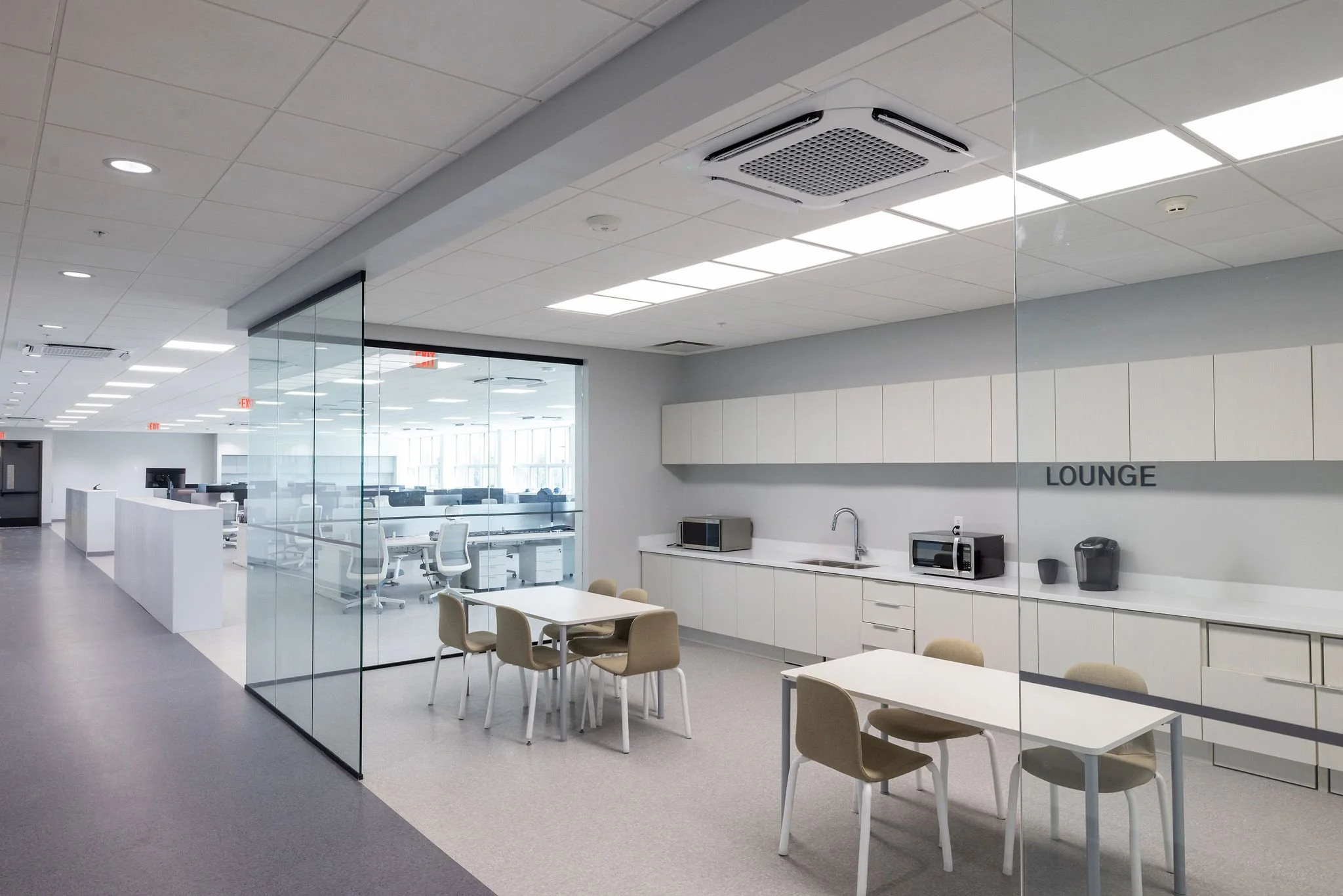Modern office lounge area with tables, chairs, kitchen appliances, and glass walls, illuminated by overhead fluorescent lights.