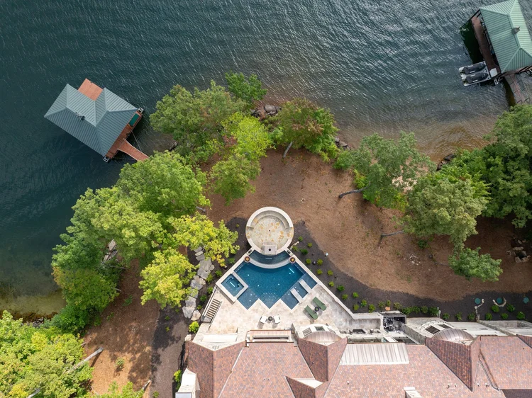 Aerial view of a backyard with a swimming pool, surrounding patio, trees, and adjacent to a lake with two docked boats.