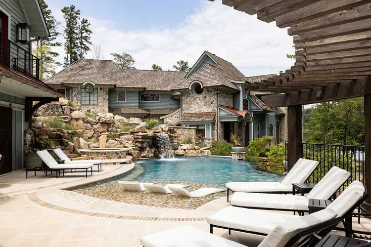 Luxury house with stone and wood exterior, a waterfall flowing into a swimming pool, surrounded by lounge chairs, with a landscaped garden and trees in the background.