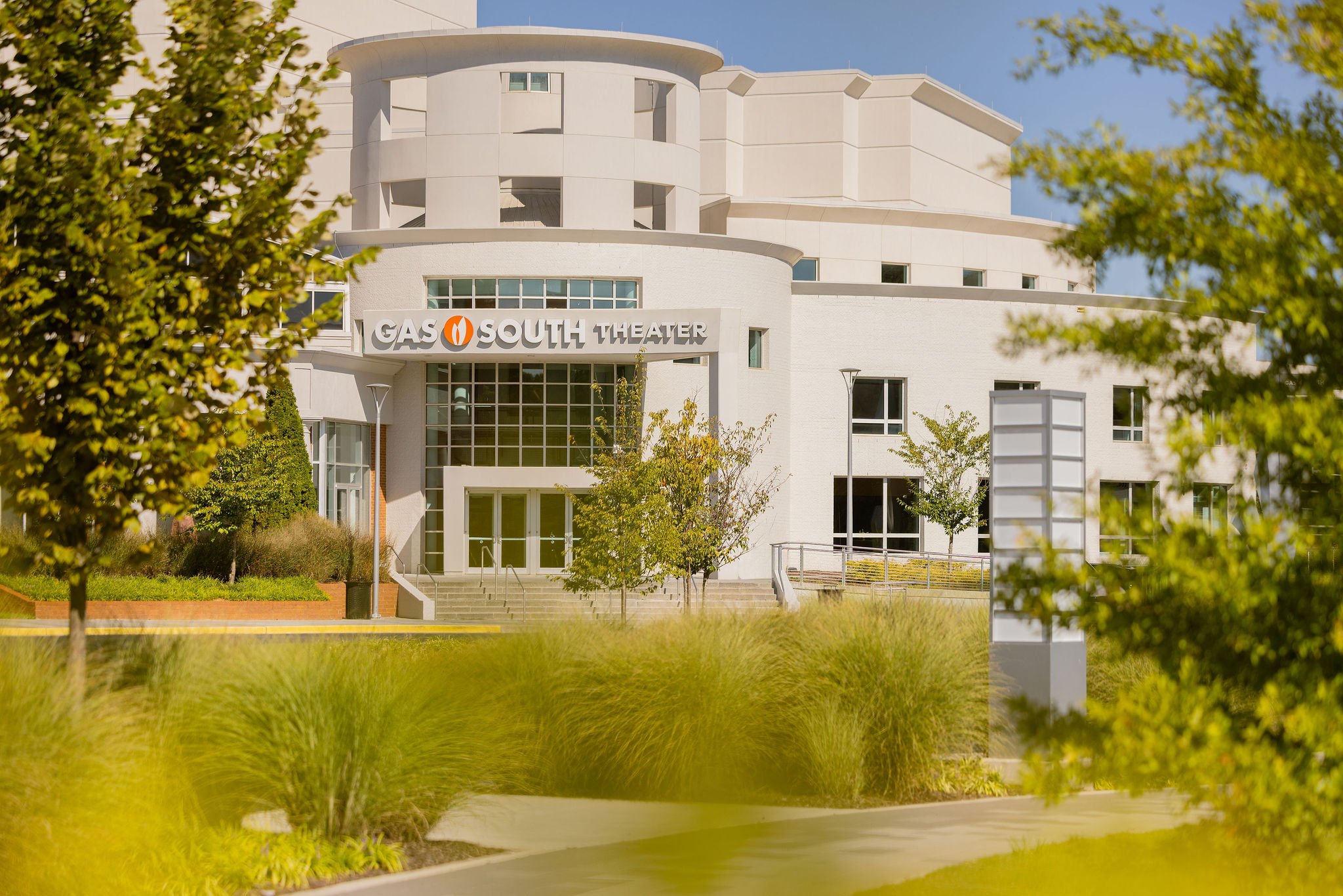 Exterior of the Gas South Theater building with a white curved facade, glass entrance, and the sign reading 'Gas South Theater' on the front. Green trees and plants partially frame the view amidst a bright, sunny day.