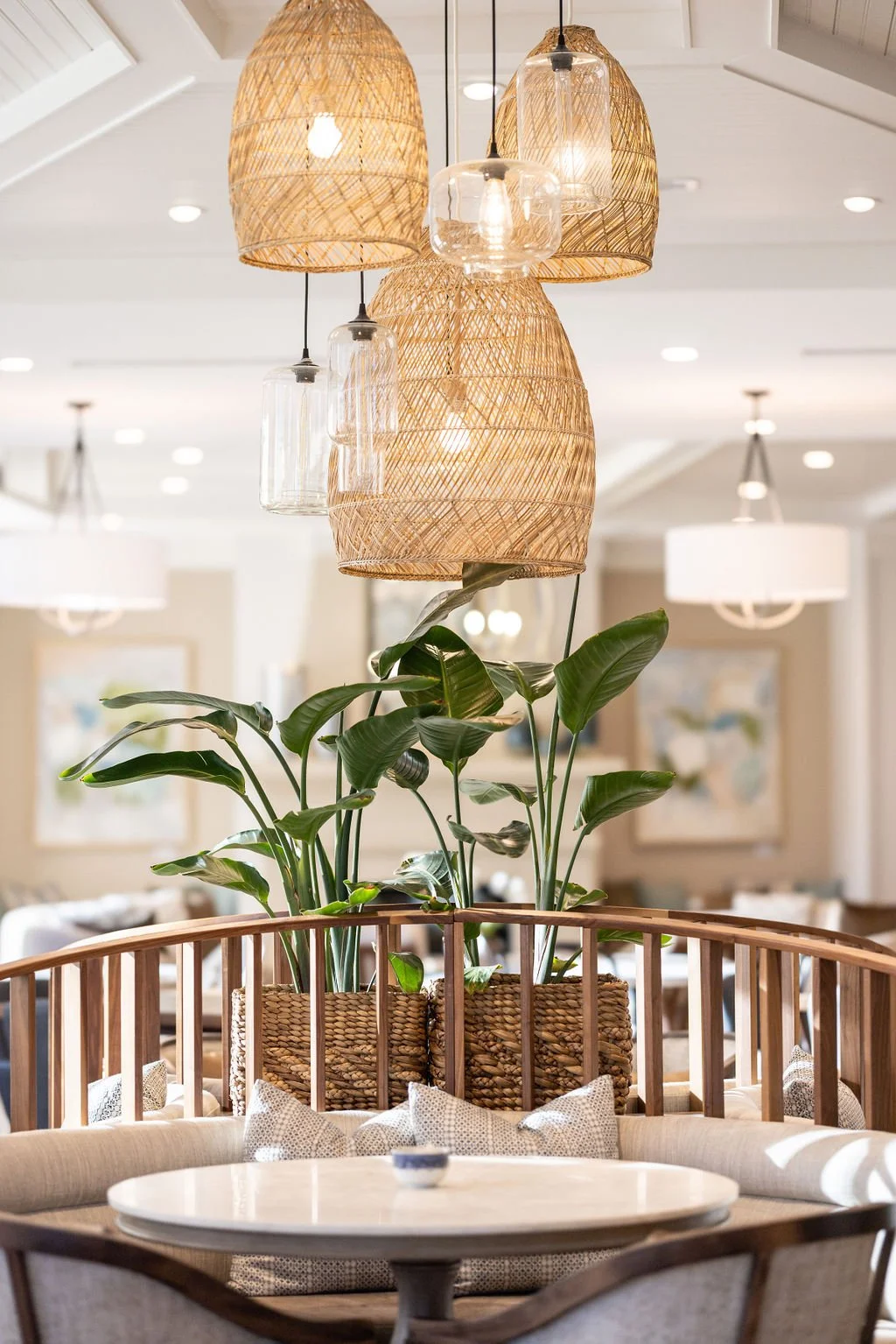 Interior of a modern dining area with hanging wicker and glass pendant lights, a large leafy plant in a woven basket, and a round table with upholstered seating.