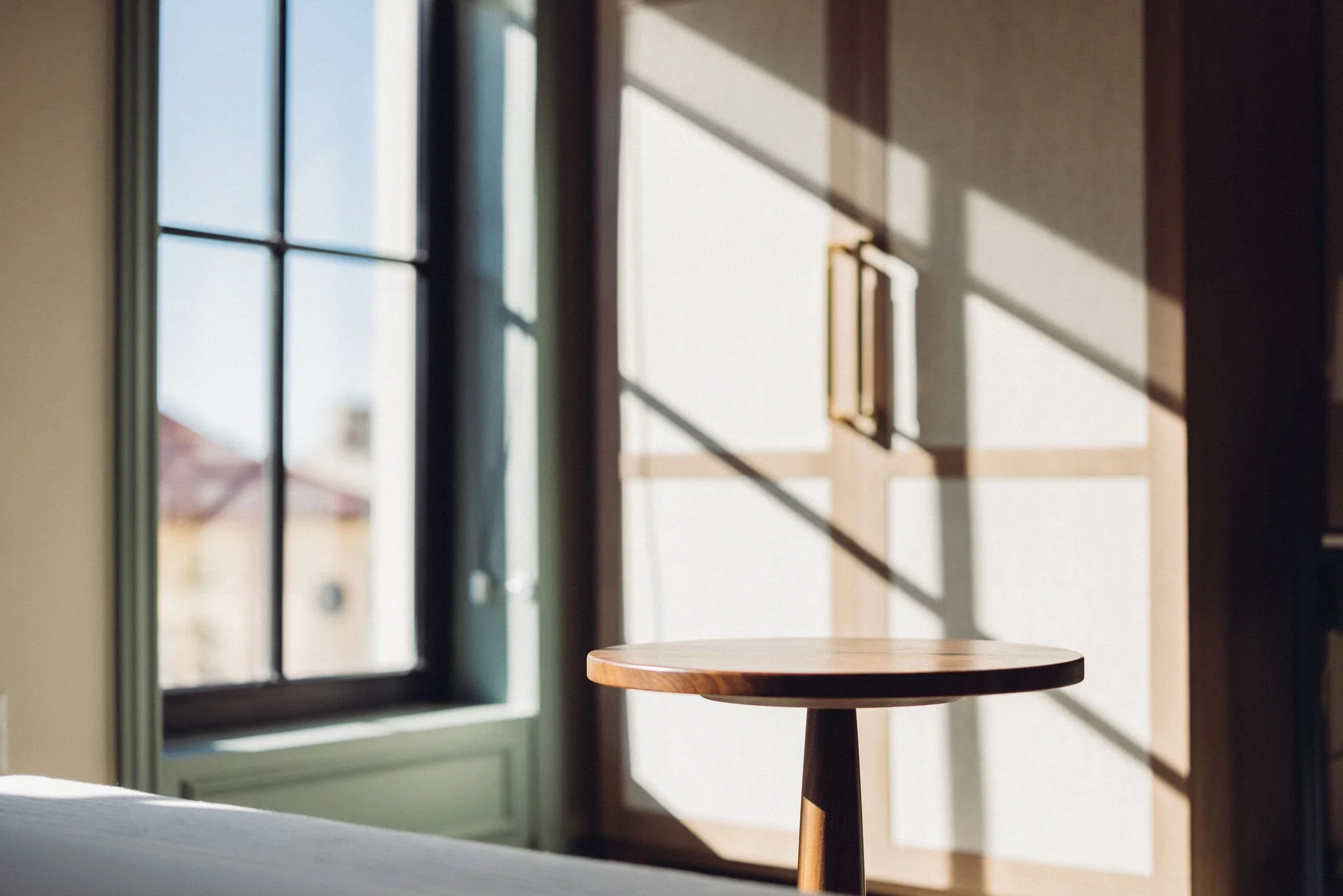 A small round wooden table next to a window with green framing. Sunlight casts shadows on a white wall behind the table.
