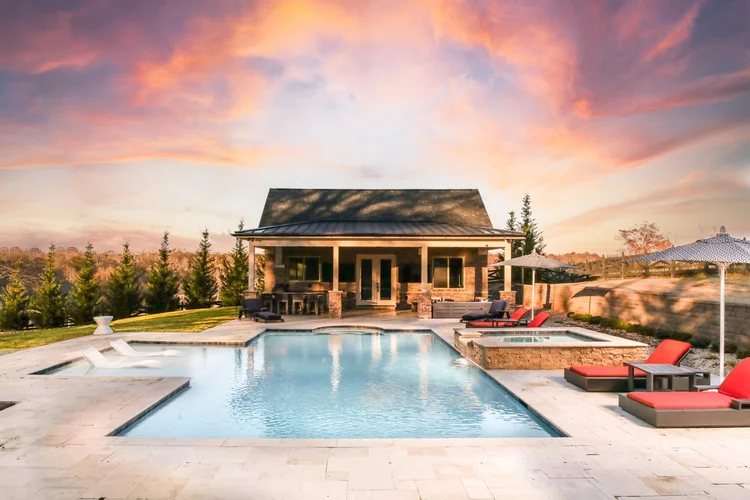 A backyard featuring a swimming pool with lounge chairs, a hot tub, a patio with outdoor furniture, and a house with a porch, under a colorful sunset sky.