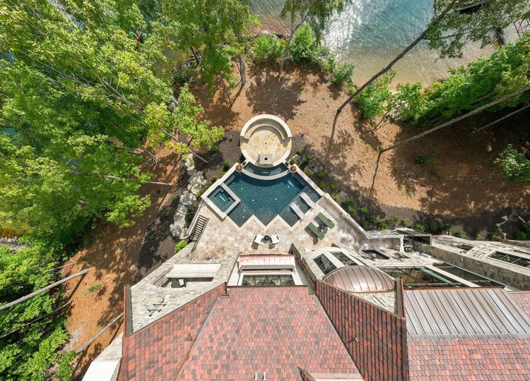 Aerial view of a backyard with a pool, hot tub, outdoor seating, and a gazebo, surrounded by tall green trees and a lake.