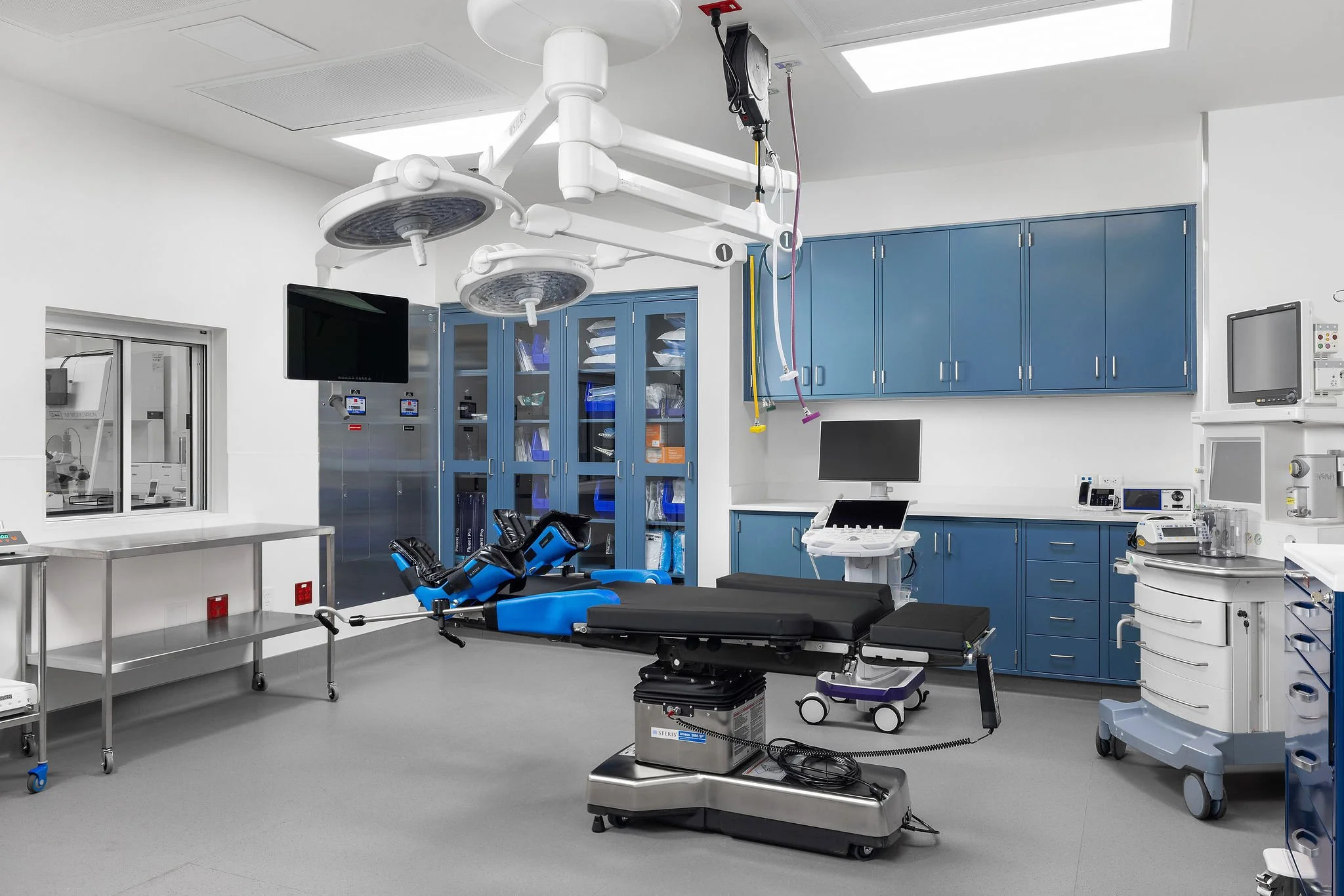A modern operating room with medical equipment including a stretcher with blue side supports, surgical lights hanging from above, blue cabinets on the wall, a monitoring screen, and various medical devices well-organized for surgery.