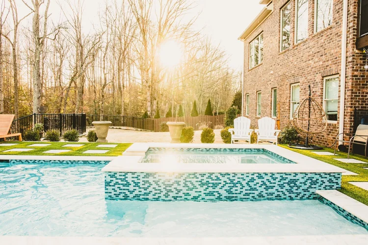Backyard with a swimming pool and hot tub, patio chairs, and a brick house with large windows under a sunny sky with leafless trees in the background.