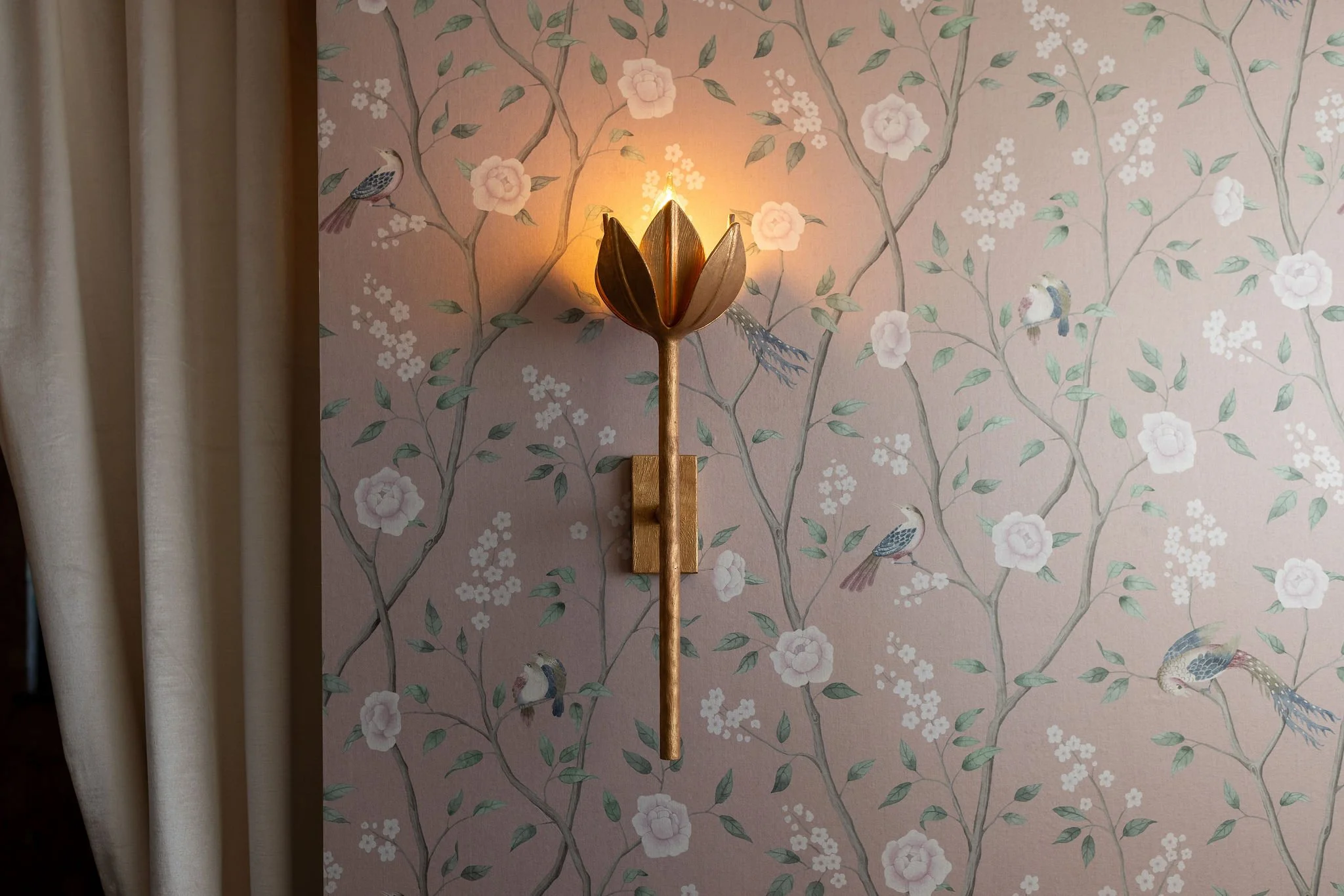 A wall sconce in the shape of a flower mounted on a pink wallpapered wall with a floral and bird pattern.