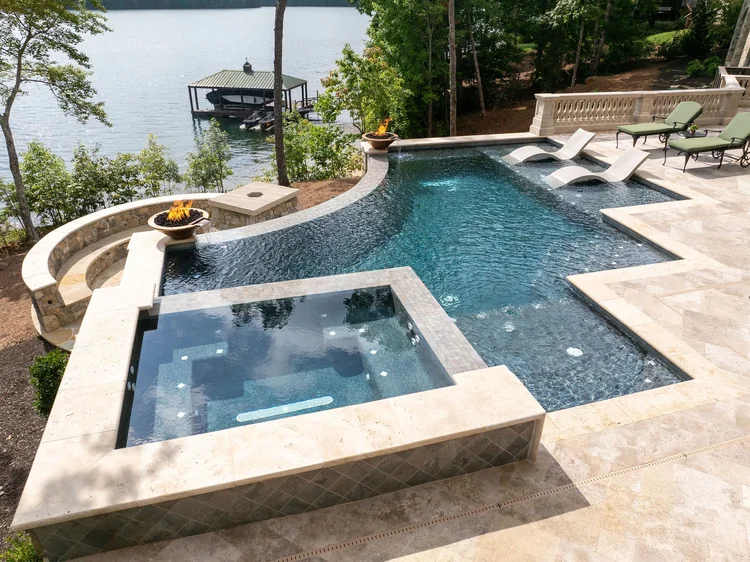 Backyard swimming pool with attached hot tub, lounge chairs, fire pits, and a lake in the background.