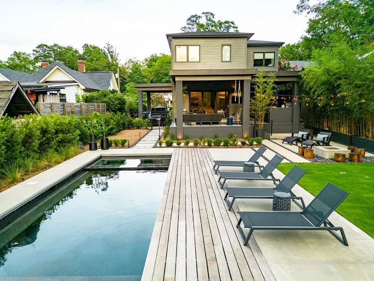 Modern backyard with swimming pool, lounge chairs, wooden deck, grassy area, and a two-story house in the background.