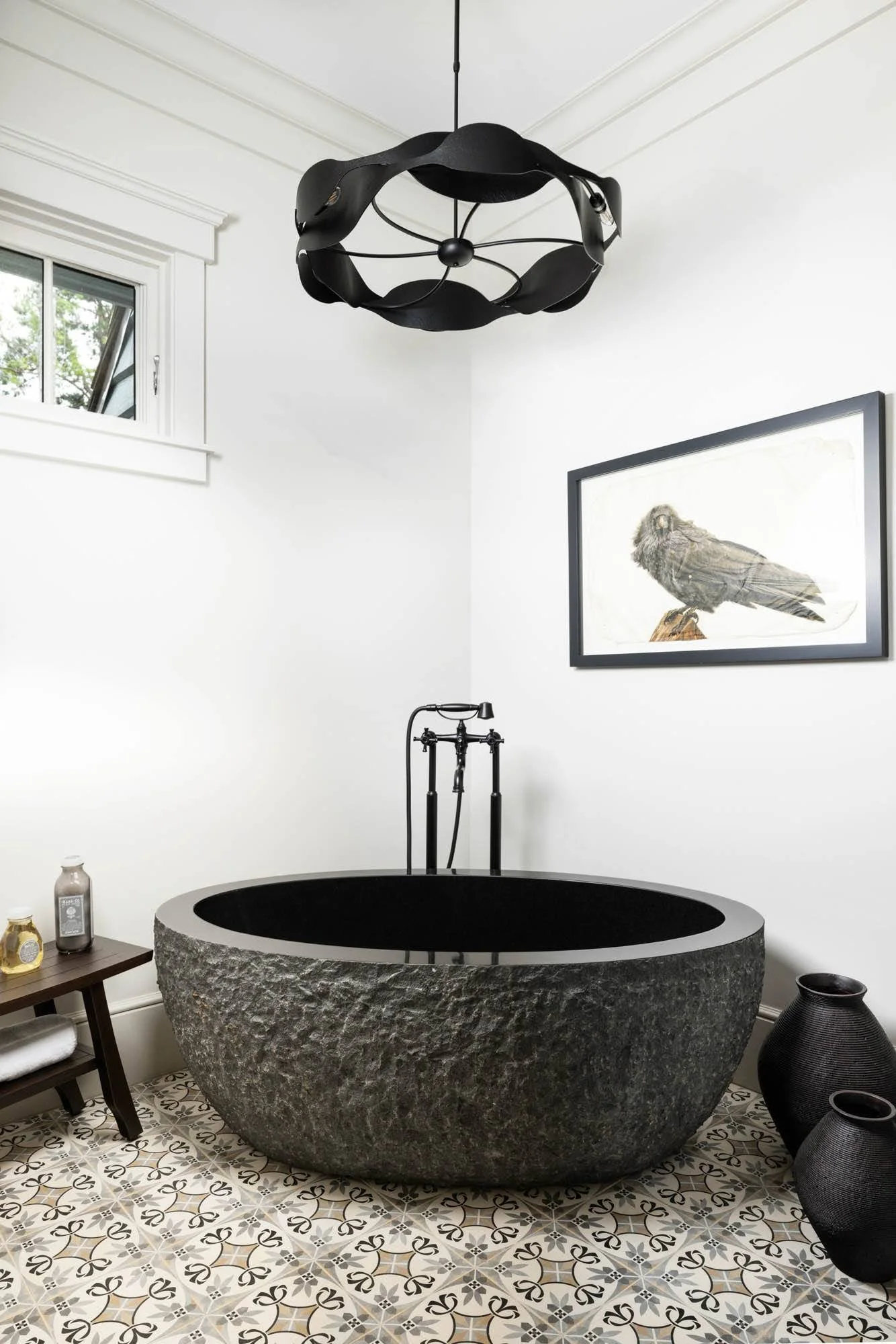 Modern bathroom with a large, textured black stone bathtub, a black ceiling light fixture, a framed bird illustration on the wall, and decorative black vases on the floor.