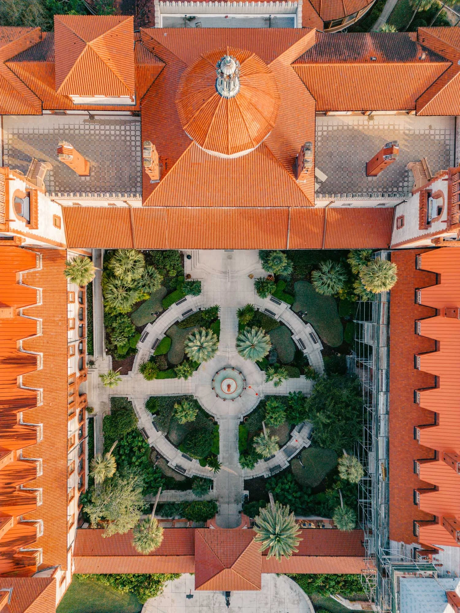 An aerial view of a large building with a red-tiled roof, surrounded by a courtyard with trees and benches. The building has a central dome and symmetrical wings.
