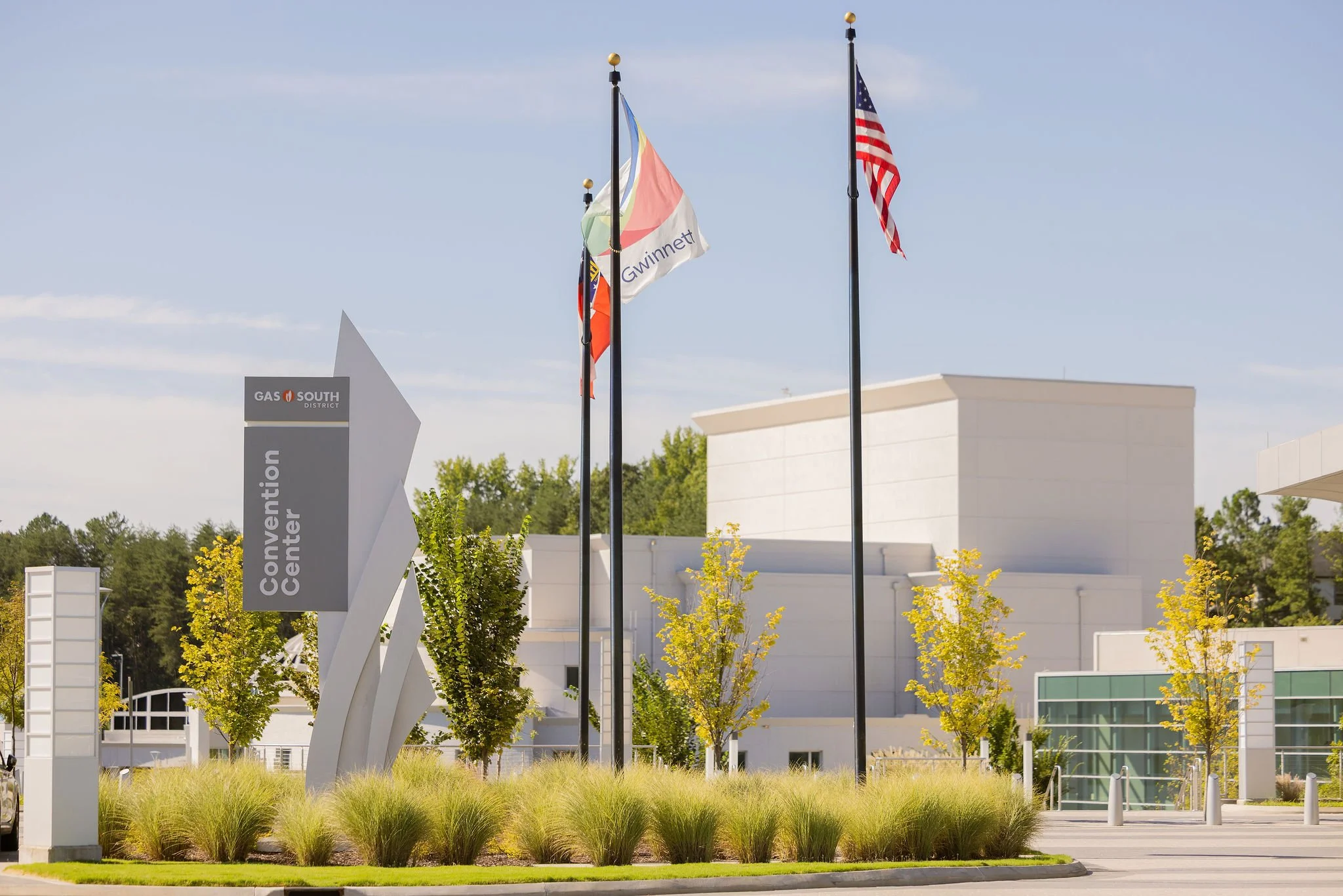 Flags of the United States and Gwinnett County, Georgia, flying beside a sign, with modern white buildings and trees in the background.