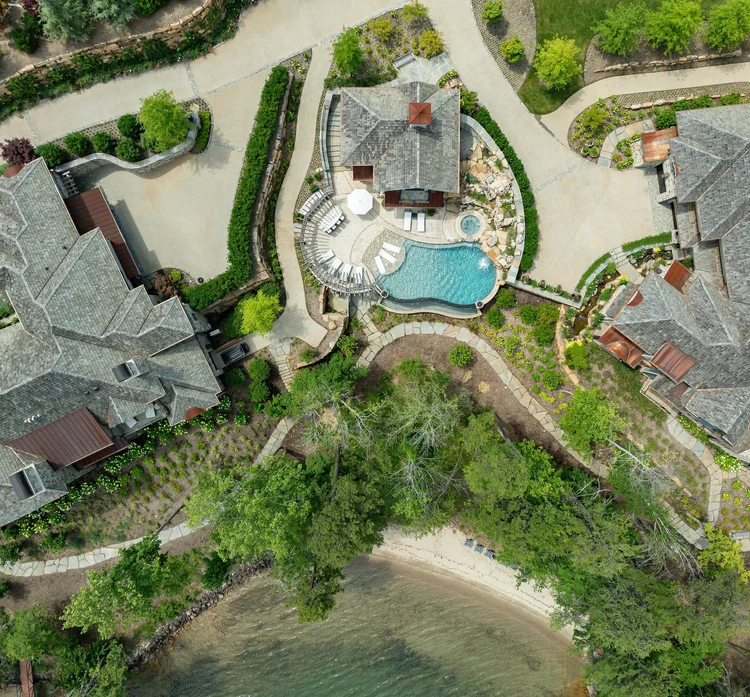Aerial view of a residential neighborhood with houses, landscaped gardens, a swimming pool, and a shoreline with trees and sandy beach.
