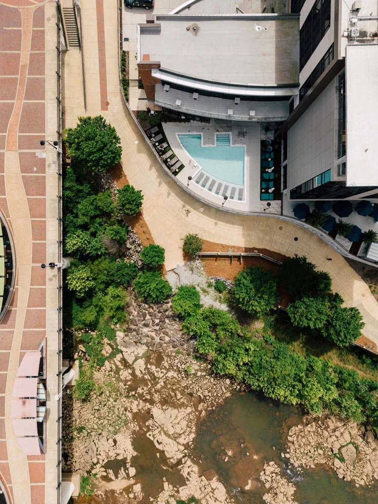 An aerial view of an urban area showing a rooftop pool, a walking path, trees, and a small body of water with rocks and plants.