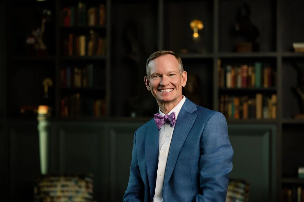 Allen Clardy standing in a blue blazer and purple bow tie in front of dark library shelves