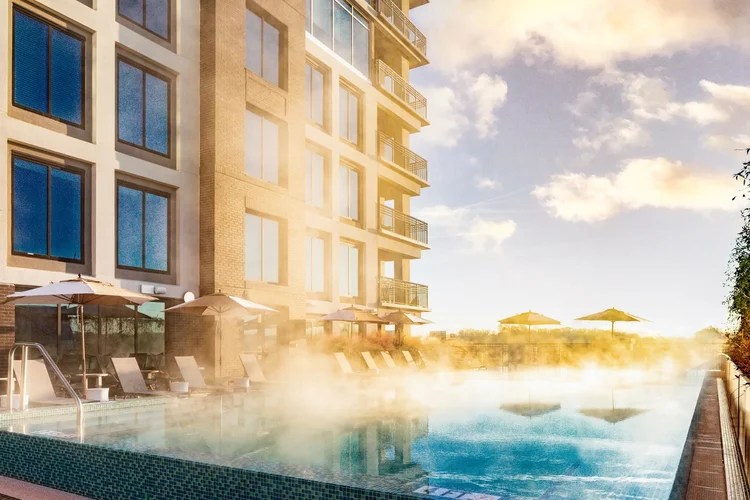 A poolside view of a modern apartment building with balconies, lounge chairs, and umbrellas, with steam rising from the pool on a sunny day.