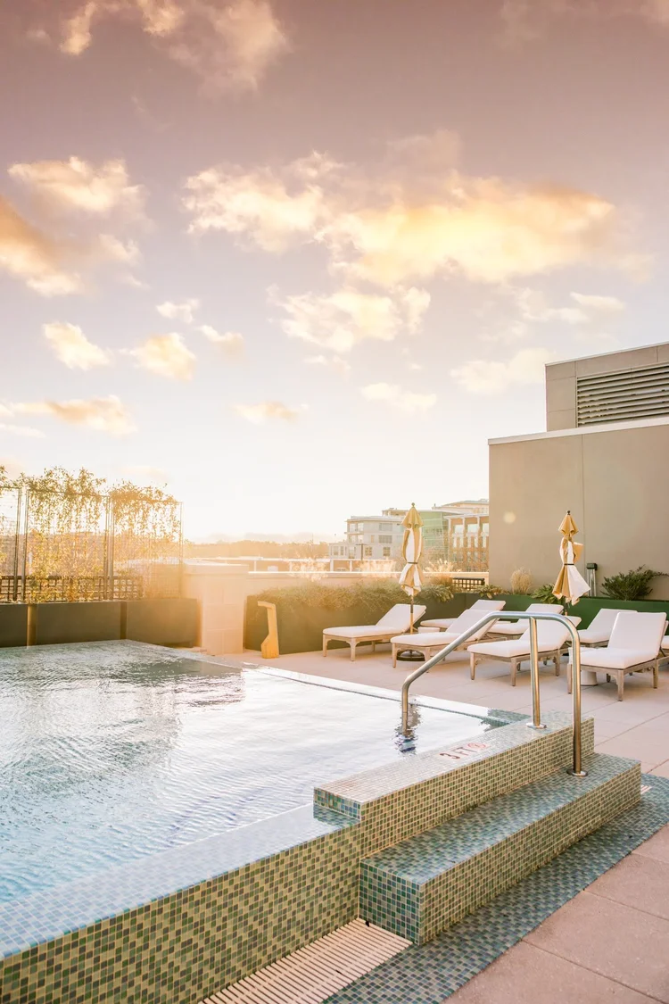 Sunset over a rooftop pool with lounge chairs and closed umbrellas, city skyline in the background.