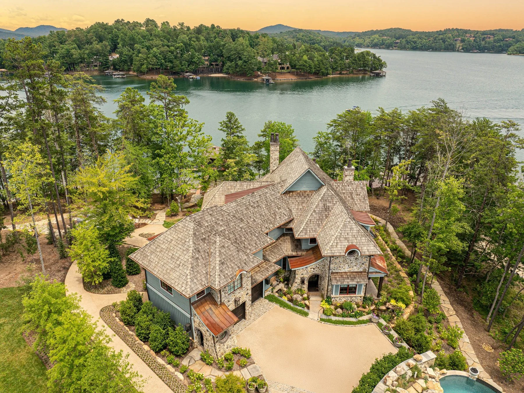 A large house with a tan tiled roof, stone and wood exterior, located on a spacious property with a stone driveway and lush landscaping, overlooking a lake with trees and distant hills in the background.